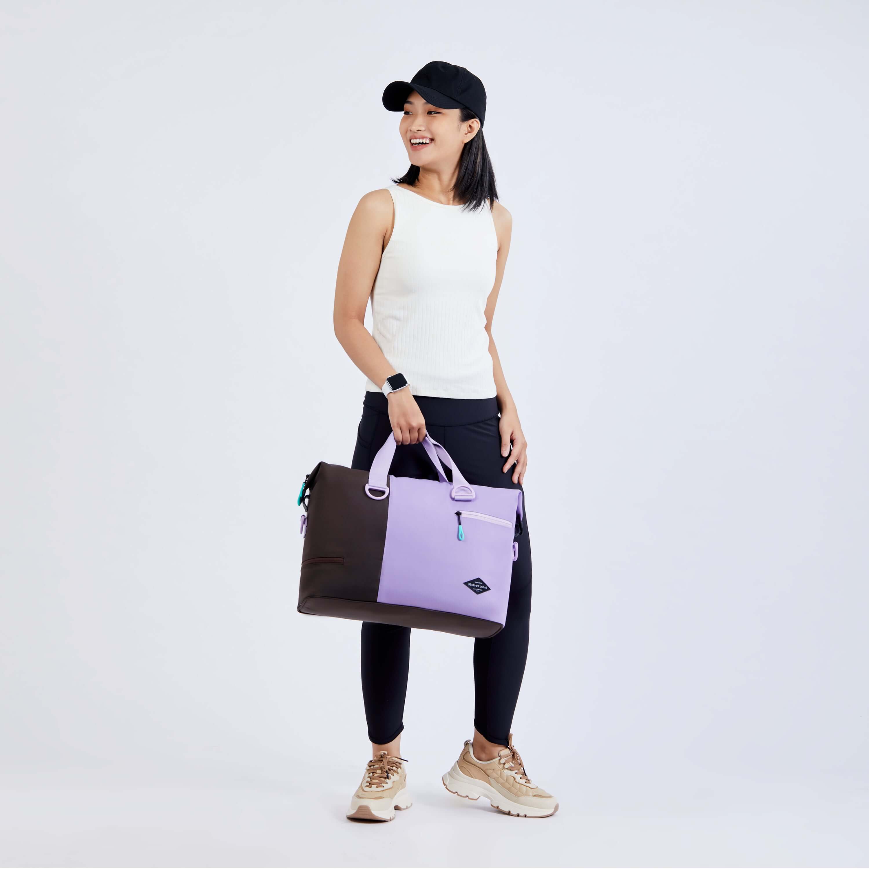 Full body view of a dark haired model facing the camera and smiling over her right shoulder. She is wearing a black ball cap, white tank top, black leggings and sneakers. She carries Sherpani bag, the Sola in Lavender, by the tote handles. 