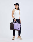 Full body view of a dark haired model facing the camera and smiling over her right shoulder. She is wearing a black ball cap, white tank top, black leggings and sneakers. She carries Sherpani bag, the Sola in Lavender, by the tote handles.