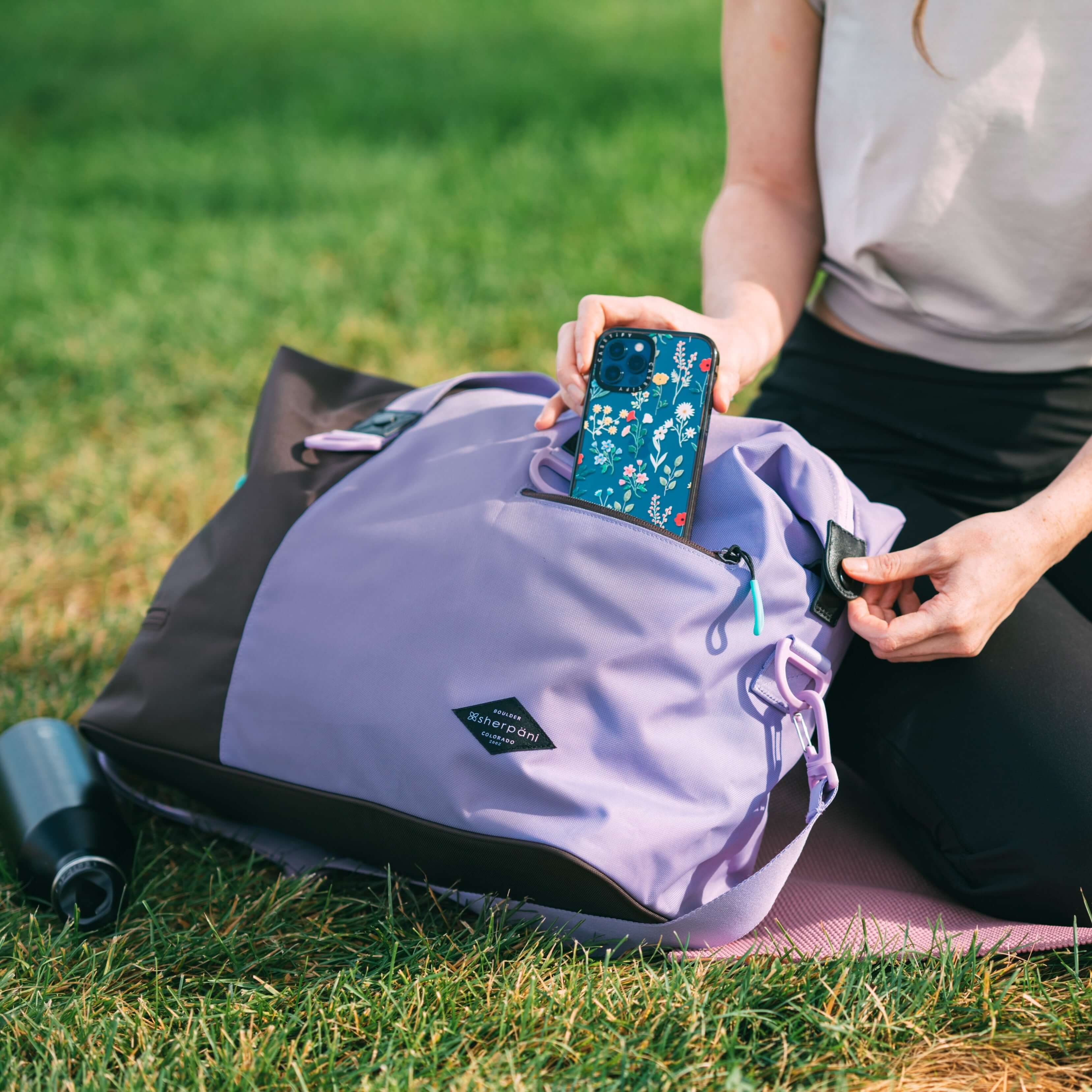Close up view of a woman sitting outside in a park on a yoga mat. She is taking her phone out of the external zipper pocket of Sherpani bag, the Sola in Lavender. 