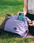 Close up view of a woman sitting outside in a park on a yoga mat. She is taking her phone out of the external zipper pocket of Sherpani bag, the Sola in Lavender.
