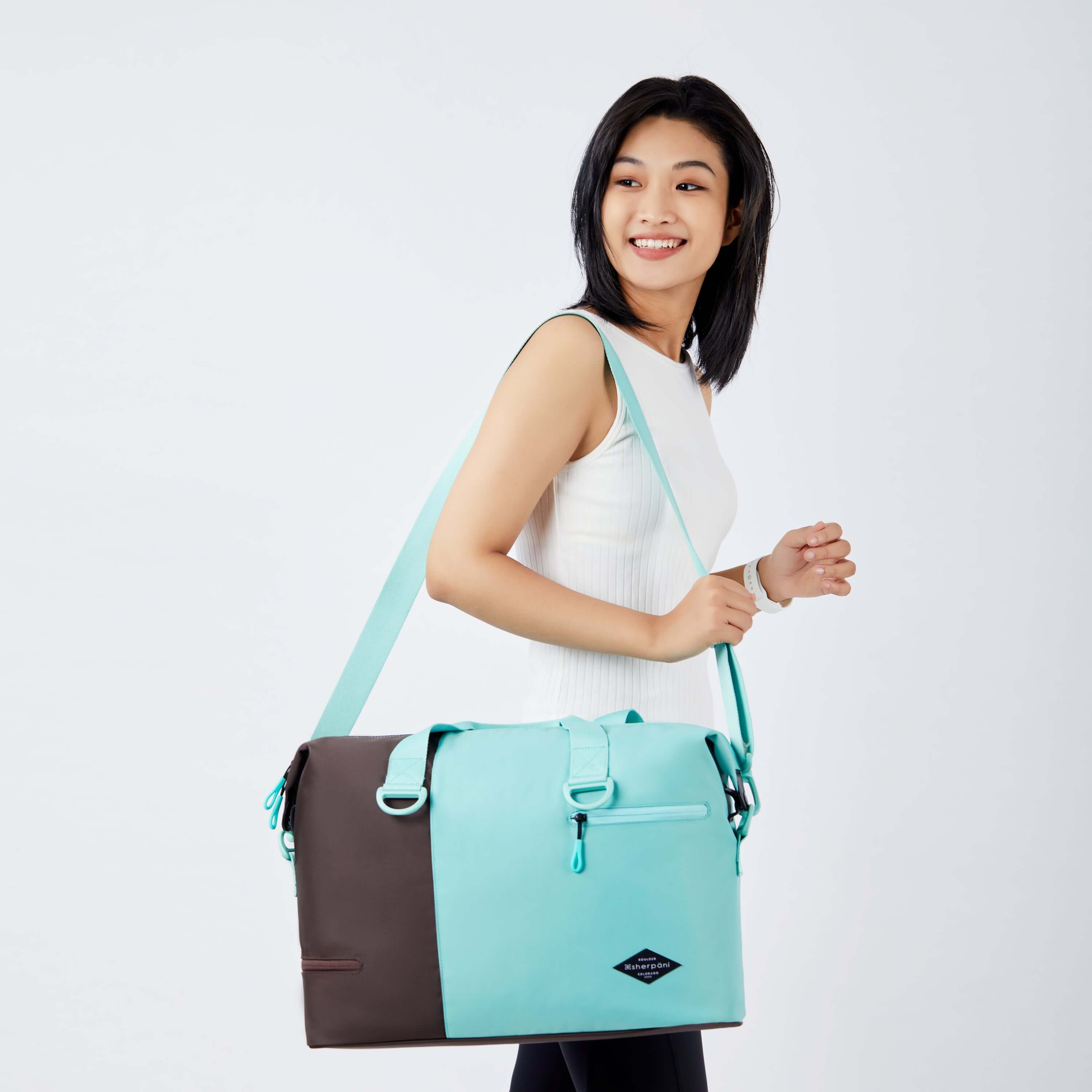 Close up view of a dark haired model facing the side and smiling over her right shoulder. She is wearing a white tank top and black leggings. She carries Sherpani bag, the Sola in Seagreen, over her shoulder. 
