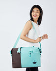 Close up view of a dark haired model facing the side and smiling over her right shoulder. She is wearing a white tank top and black leggings. She carries Sherpani bag, the Sola in Seagreen, over her shoulder.