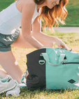 A red haired woman crouches on the grass outside. She is wearing a white tank top and athletic shorts. Sherpani bag, the Sola in Seagreen, sits on the ground in front of her. She is looking into the bag. In front of the bag a pair of sneakers lies on the grass.
