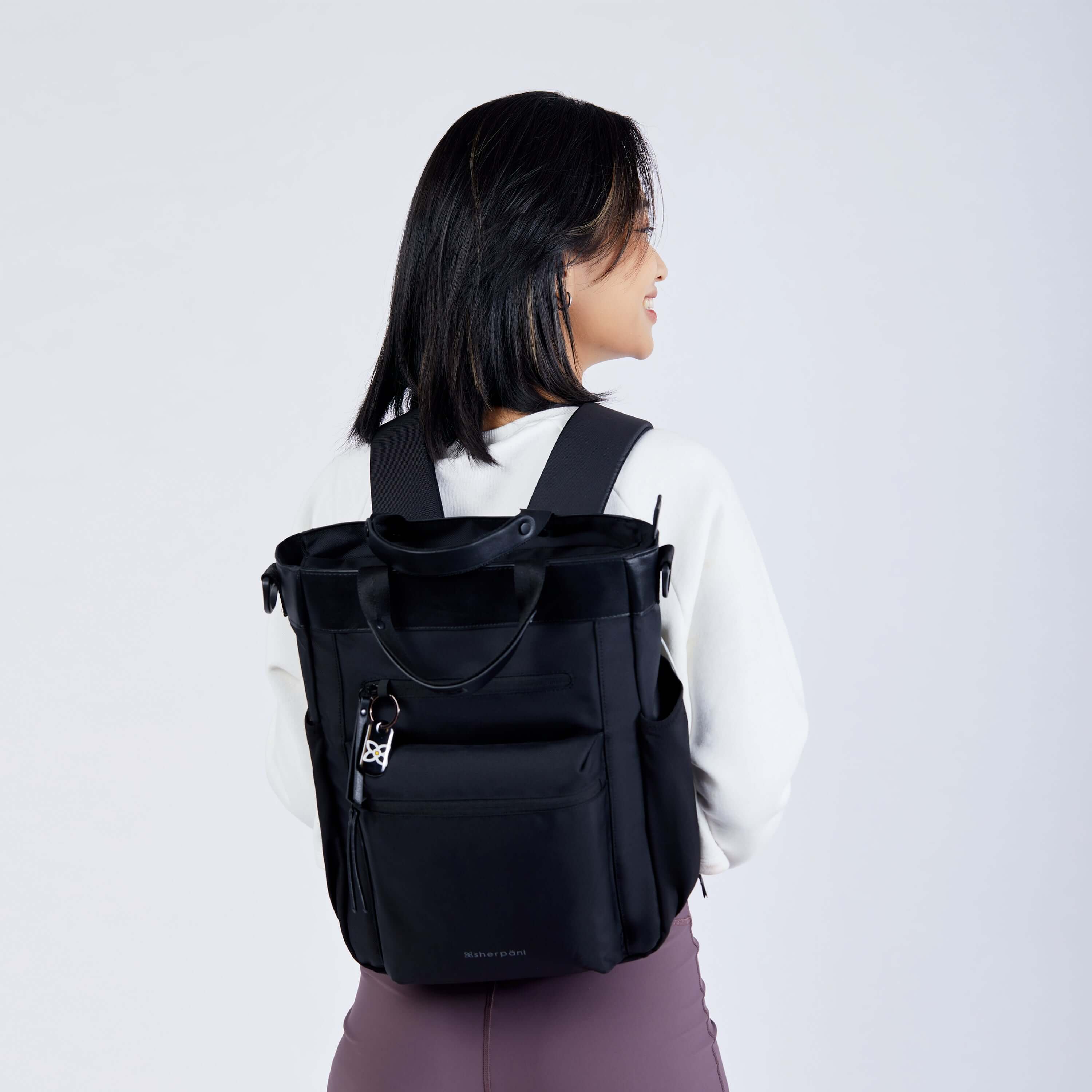 Close up view of a dark haired model facing away from the camera and smiling over her right shoulder. She is wearing a white sweatshirt, purple leggings and Sherpani’s Anti-Theft bag, the Soleil AT in Carbon, as a backpack.