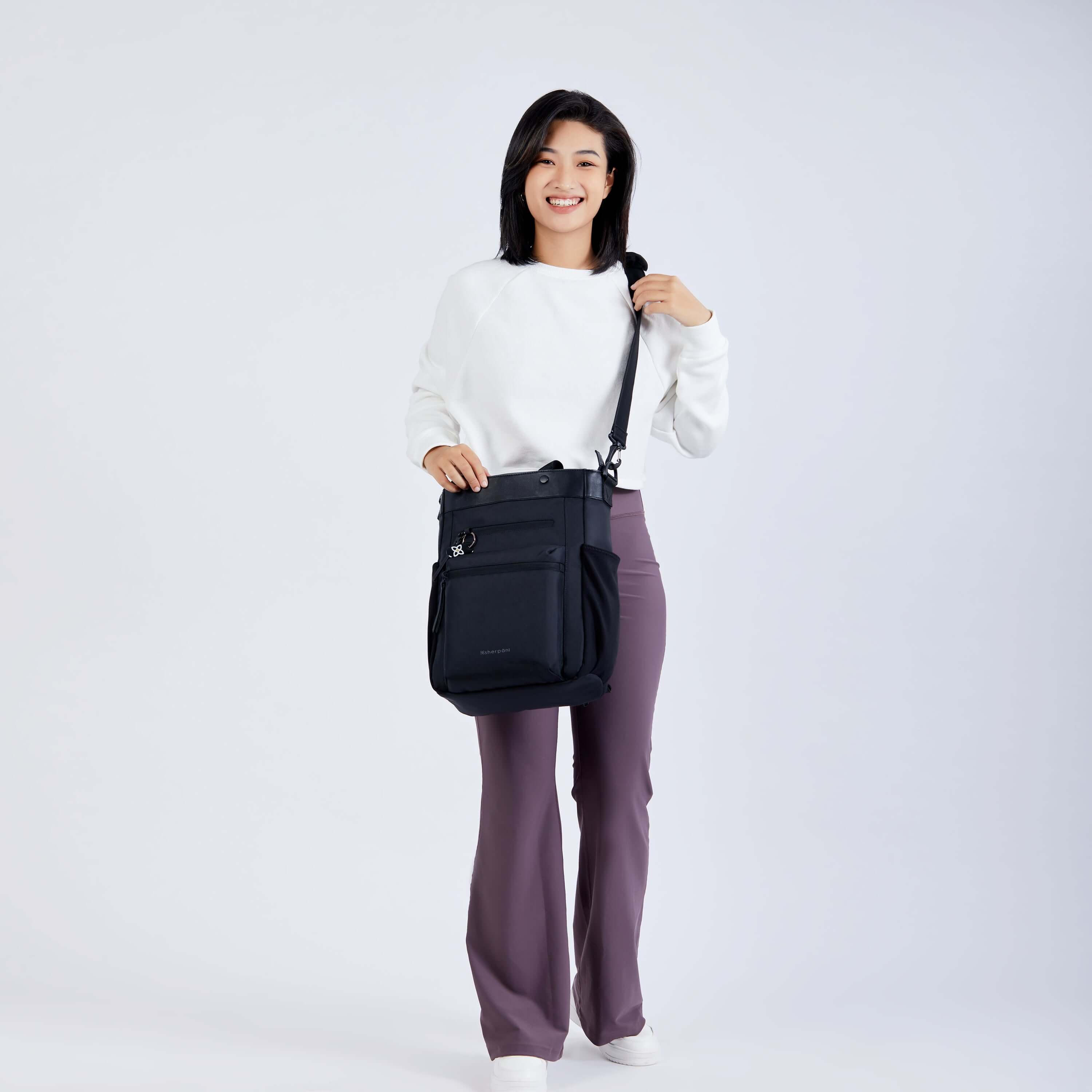 Full body view of a dark haired model facing the camera and smiling. She is wearing a white sweatshirt, purple leggings and Sherpani’s Anti-Theft bag, the Soleil AT in Carbon, as a crossbody.