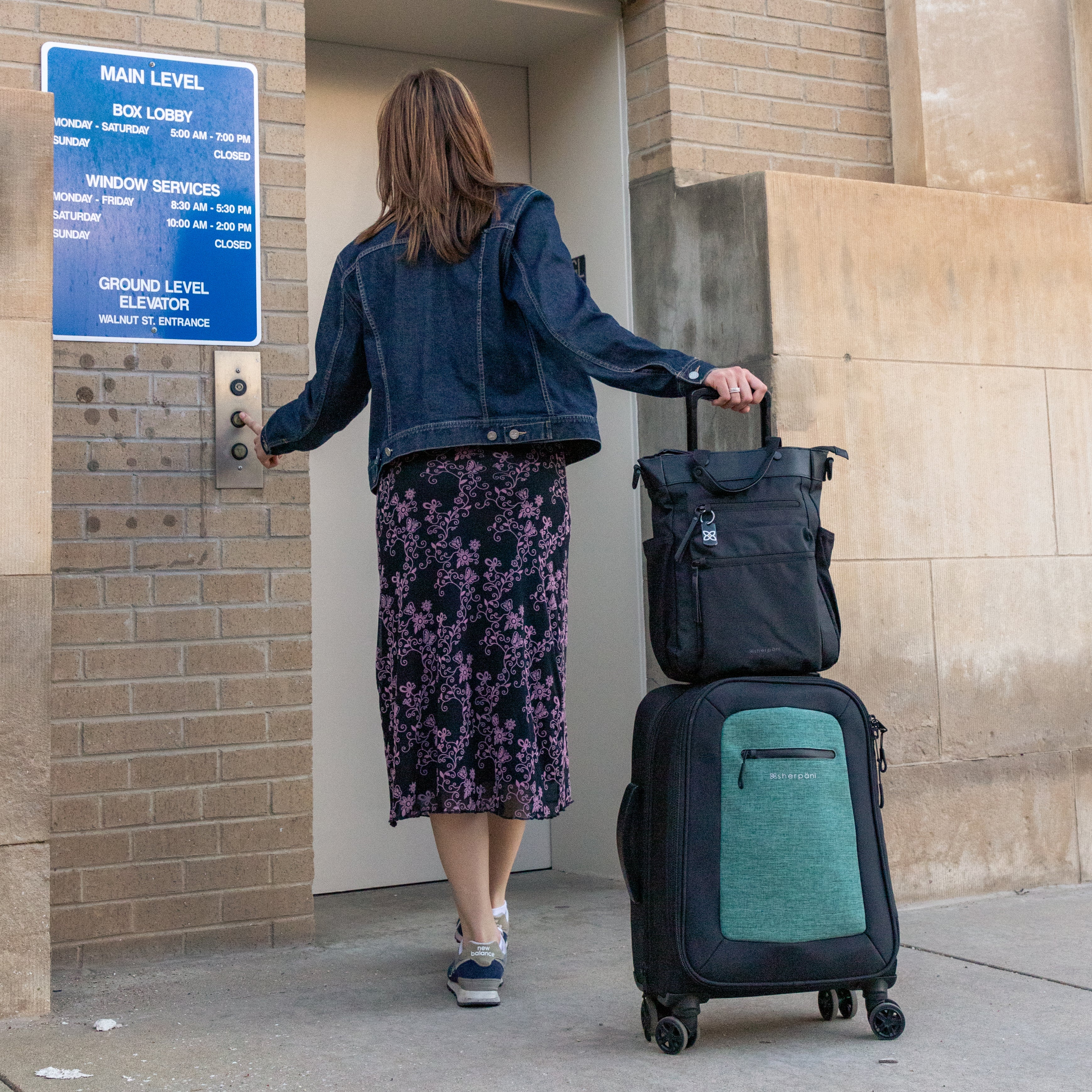 A woman pushes an elevator button in an airport parking garage. She has Sherpani Anti-Theft travel bag, the Soleil in Carbon, sitting on top of Sherpani soft-shell luggage, utilizing the luggage pass through feature. 