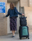 A woman pushes an elevator button in an airport parking garage. She has Sherpani Anti-Theft travel bag, the Soleil in Carbon, sitting on top of Sherpani soft-shell luggage, utilizing the luggage pass through feature.