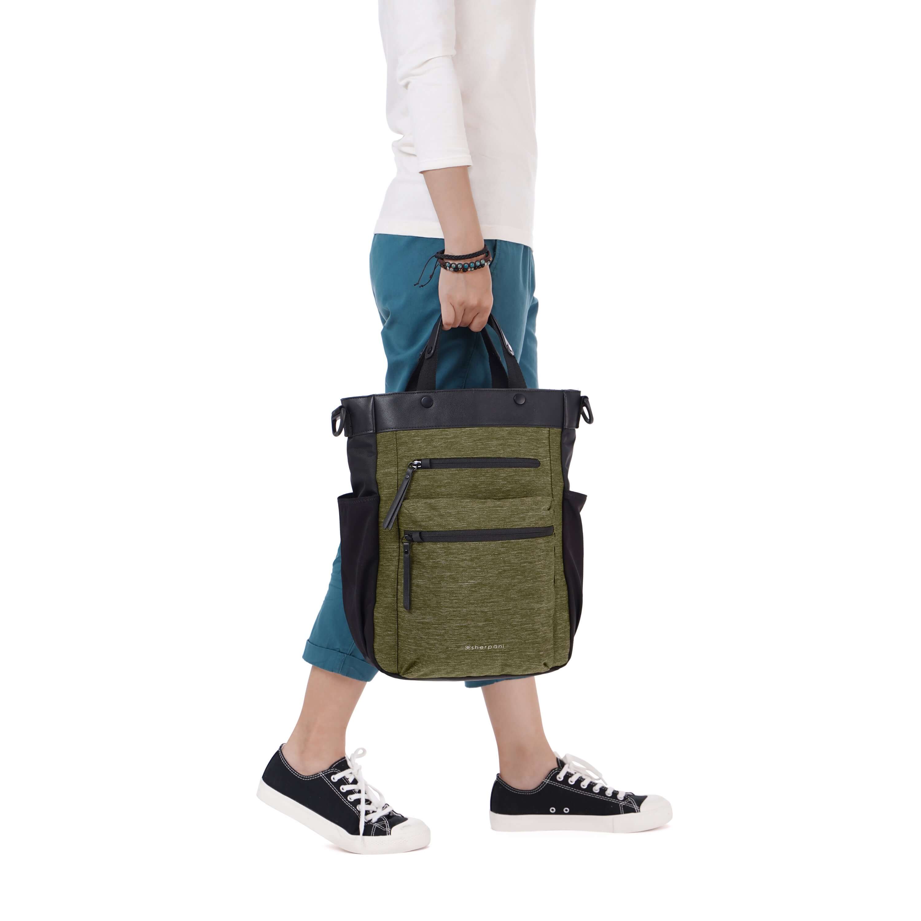 Side view of a model walking. She is wearing a white shirt and blue pants. She is carrying Sherpani’s Anti-Theft bag, the Soleil AT in Loden by the tote handles.