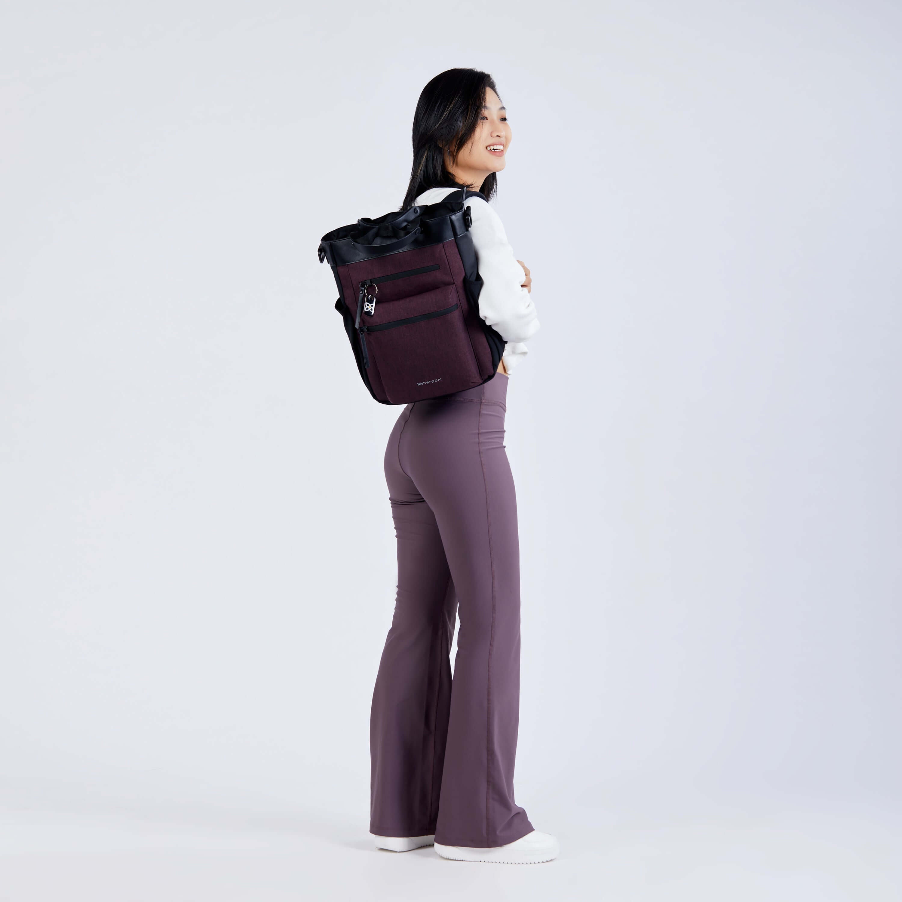 Full body view of a dark haired model facing away from the camera and smiling over her right shoulder. She is wearing a white sweatshirt, purple leggings and Sherpani’s Anti-Theft bag, the Soleil AT in Merlot, as a backpack.