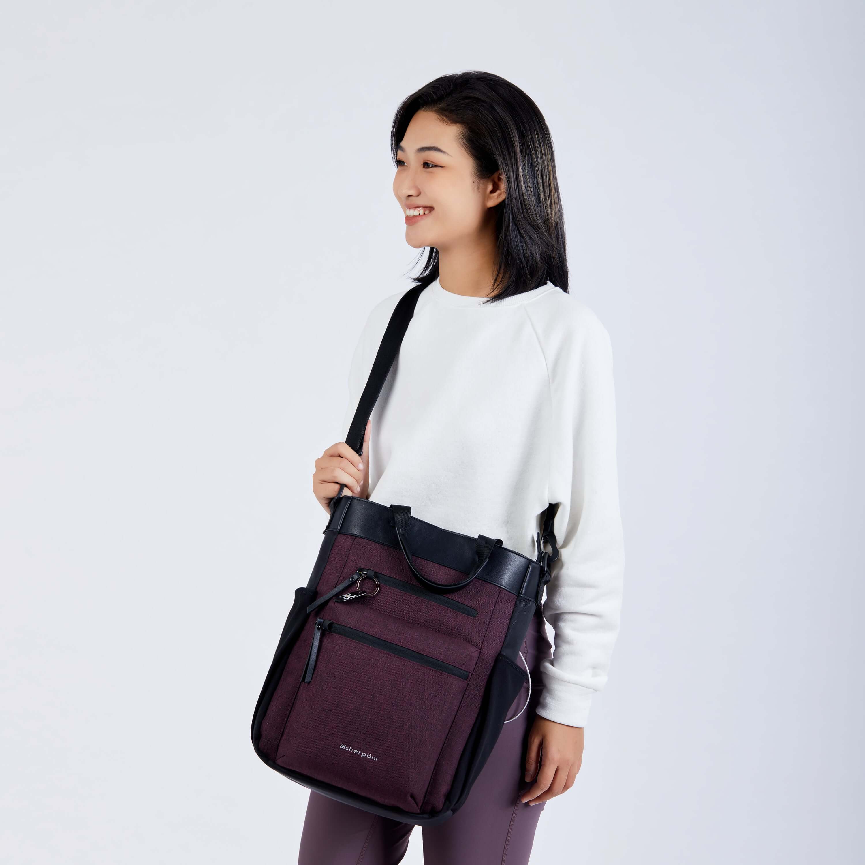Close up view of a dark haired model facing the camera and smiling. She is wearing a white sweatshirt, purple leggings, and Sherpani’s Anti-Theft bag, the Soleil AT in Merlot, as a crossbody. 