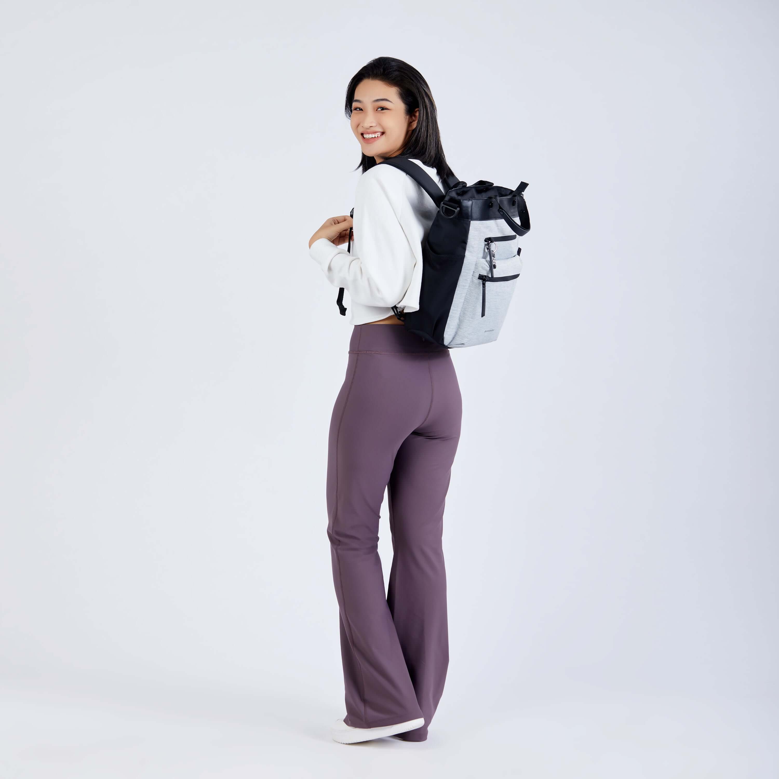 Full body view of a dark haired model facing away from the camera and smiling over her left shoulder. She is wearing a white sweatshirt, purple leggings and Sherpani’s Anti-Theft bag, the Soleil AT in Sterling, as a backpack. 