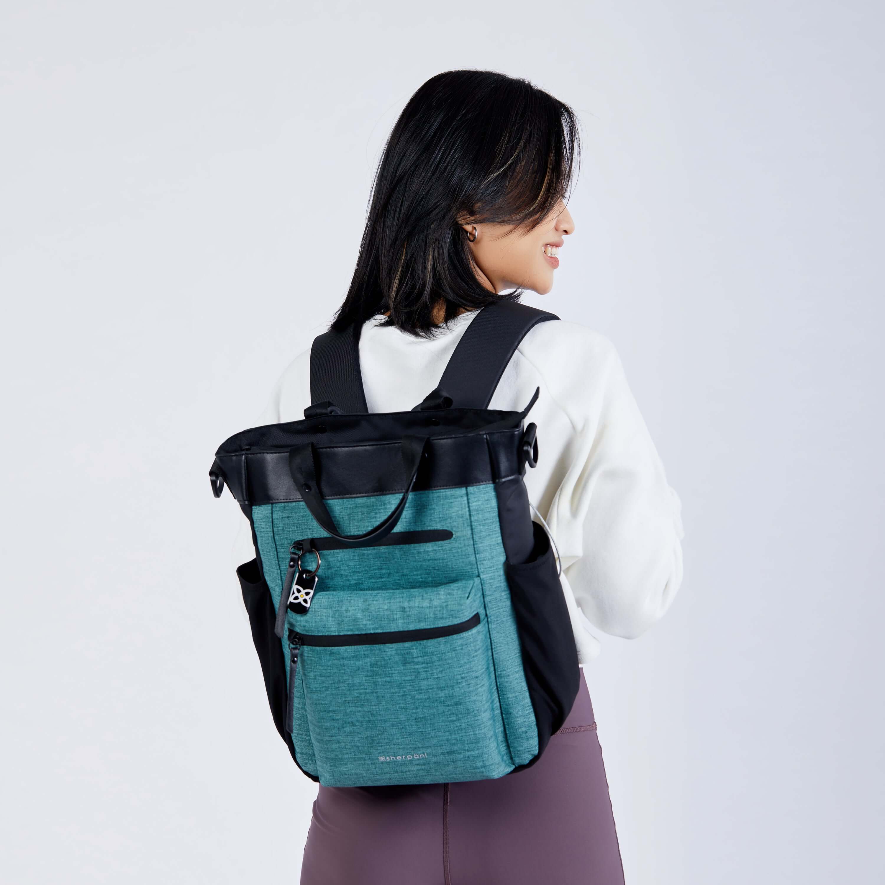 Close up view of a dark haired model facing away from the camera and smiling over her right shoulder. She is wearing a white sweatshirt, purple leggings and Sherpani’s Anti-Theft bag, the Soleil AT in Teal, as a backpack.