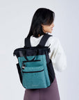 Close up view of a dark haired model facing away from the camera and smiling over her right shoulder. She is wearing a white sweatshirt, purple leggings and Sherpani’s Anti-Theft bag, the Soleil AT in Teal, as a backpack.