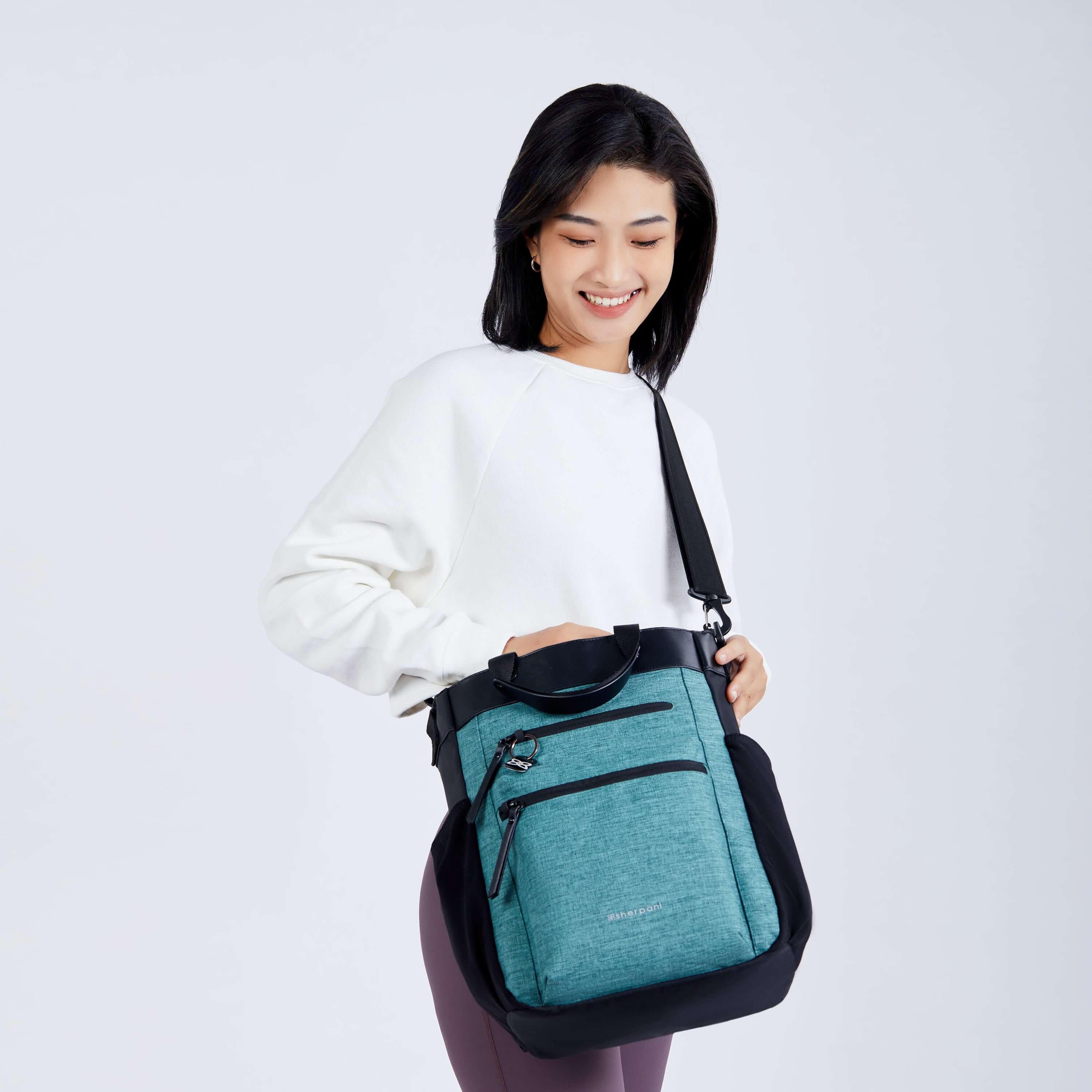 Close up view of a dark haired model facing the camera and smiling. She is wearing a white sweatshirt, purple leggings and Sherpani’s Anti-Theft bag, The Soleil AT in Teal, as a crossbody. 