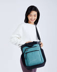Close up view of a dark haired model facing the camera and smiling. She is wearing a white sweatshirt, purple leggings and Sherpani’s Anti-Theft bag, The Soleil AT in Teal, as a crossbody.