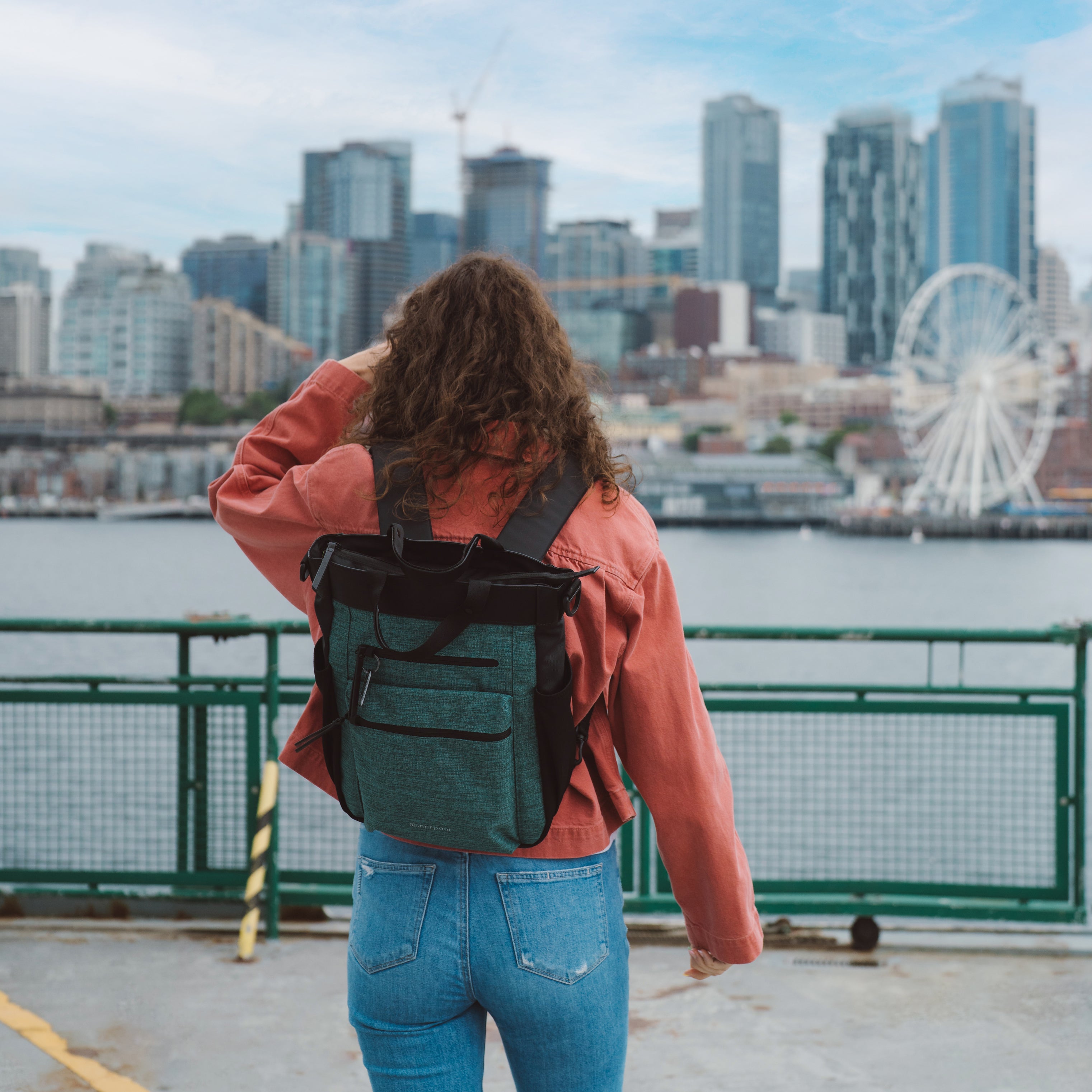 A woman stands by the waterfront and skyline of downtown Seattle. She is wearing Sherpani Anti-Theft travel bag, the Soleil in Teal, as a backpack. 