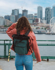 A woman stands by the waterfront and skyline of downtown Seattle. She is wearing Sherpani Anti-Theft travel bag, the Soleil in Teal, as a backpack.