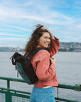 A woman stands by the sea and smiles over her shoulder. She is wearing Sherpani Anti-Theft travel bag, the Soleil in Teal, as a backpack.