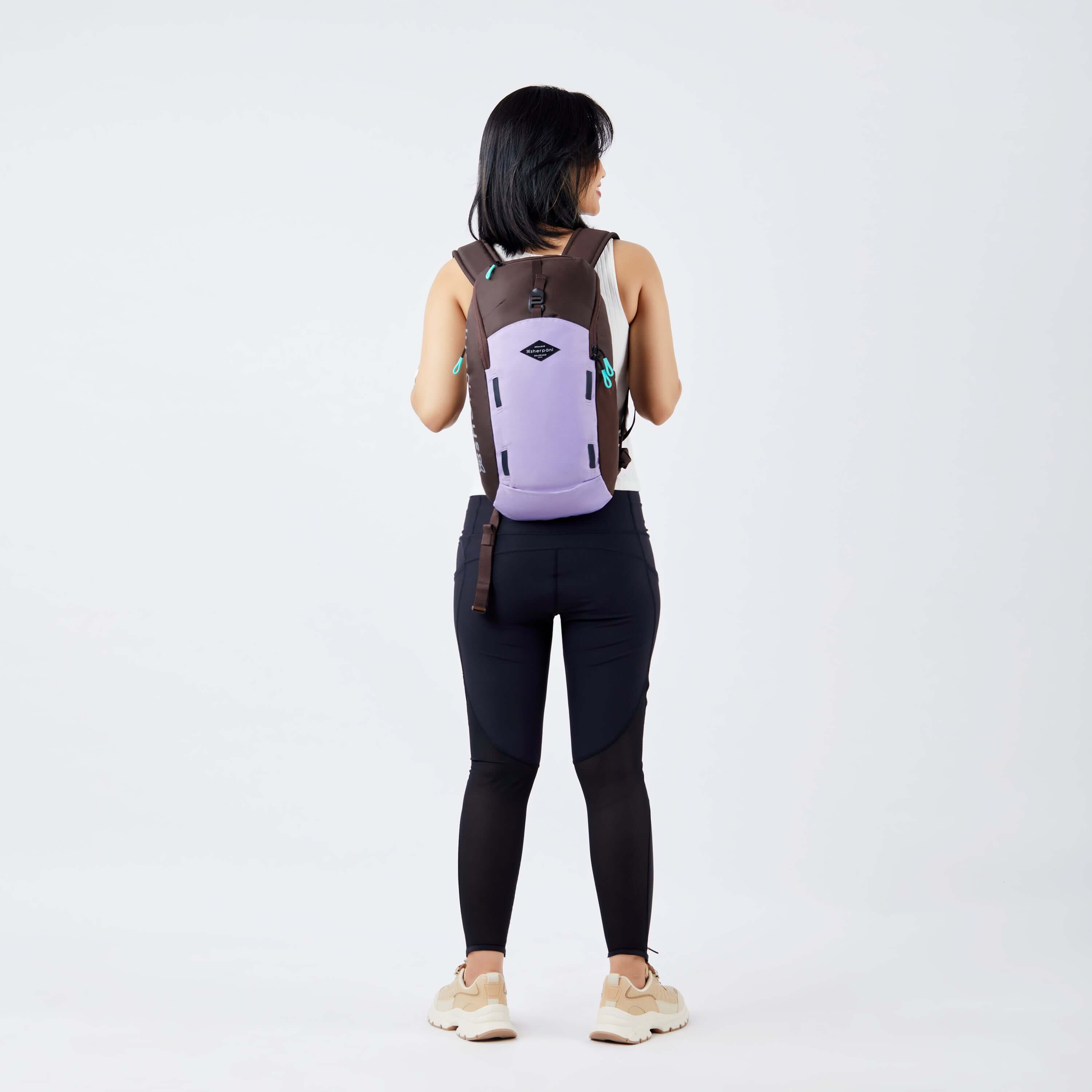 Full body view of a dark haired model facing away from the camera. She is wearing a white tank top, black leggings and sneakers. She carries Sherpani backpack, the Switch in Lavender, on her shoulders. 