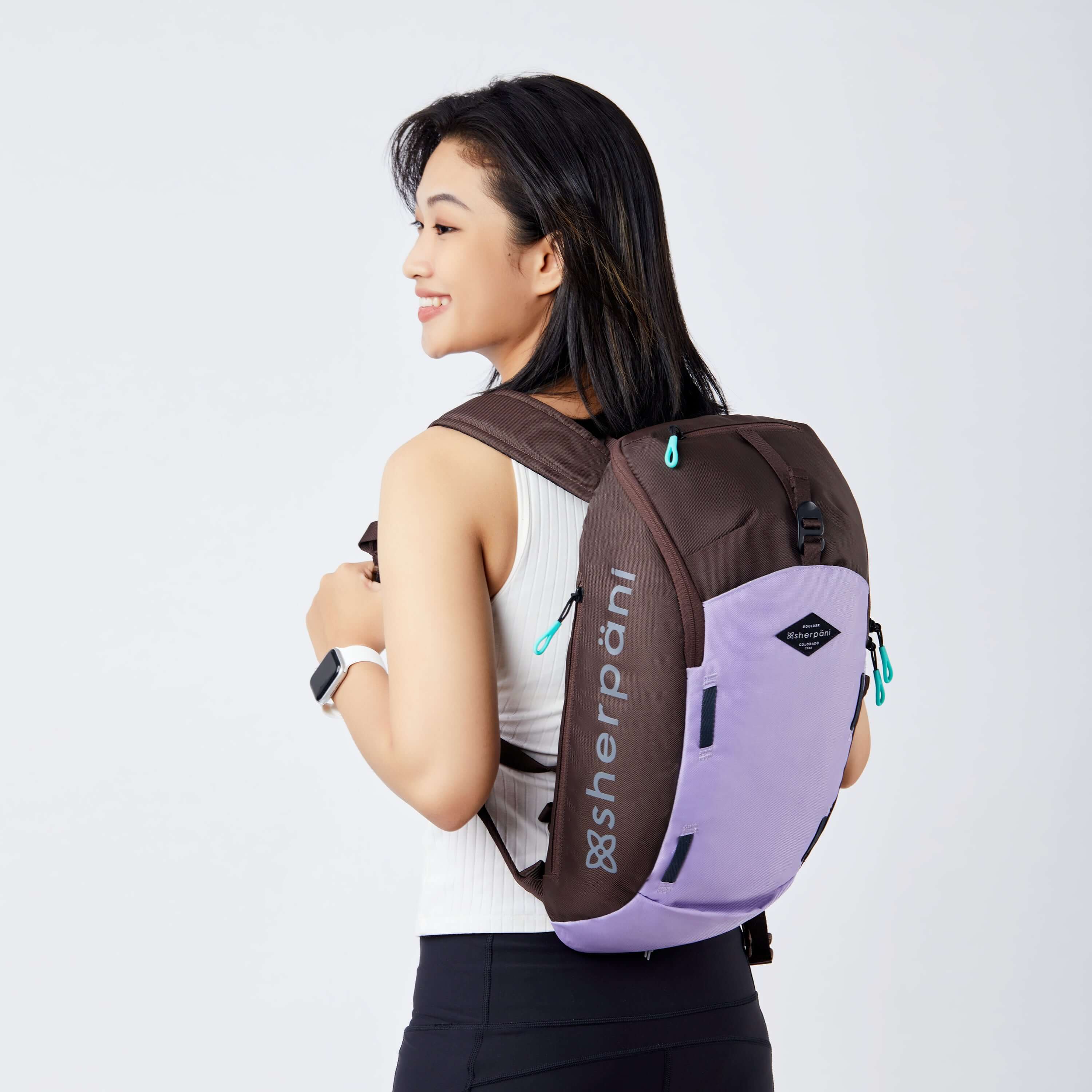 Close up view of a dark haired model facing away from the camera and smiling over her left shoulder. She is wearing a white tank top and black leggings. She carries Sherpani backpack, the Switch in Lavender, on her shoulders.