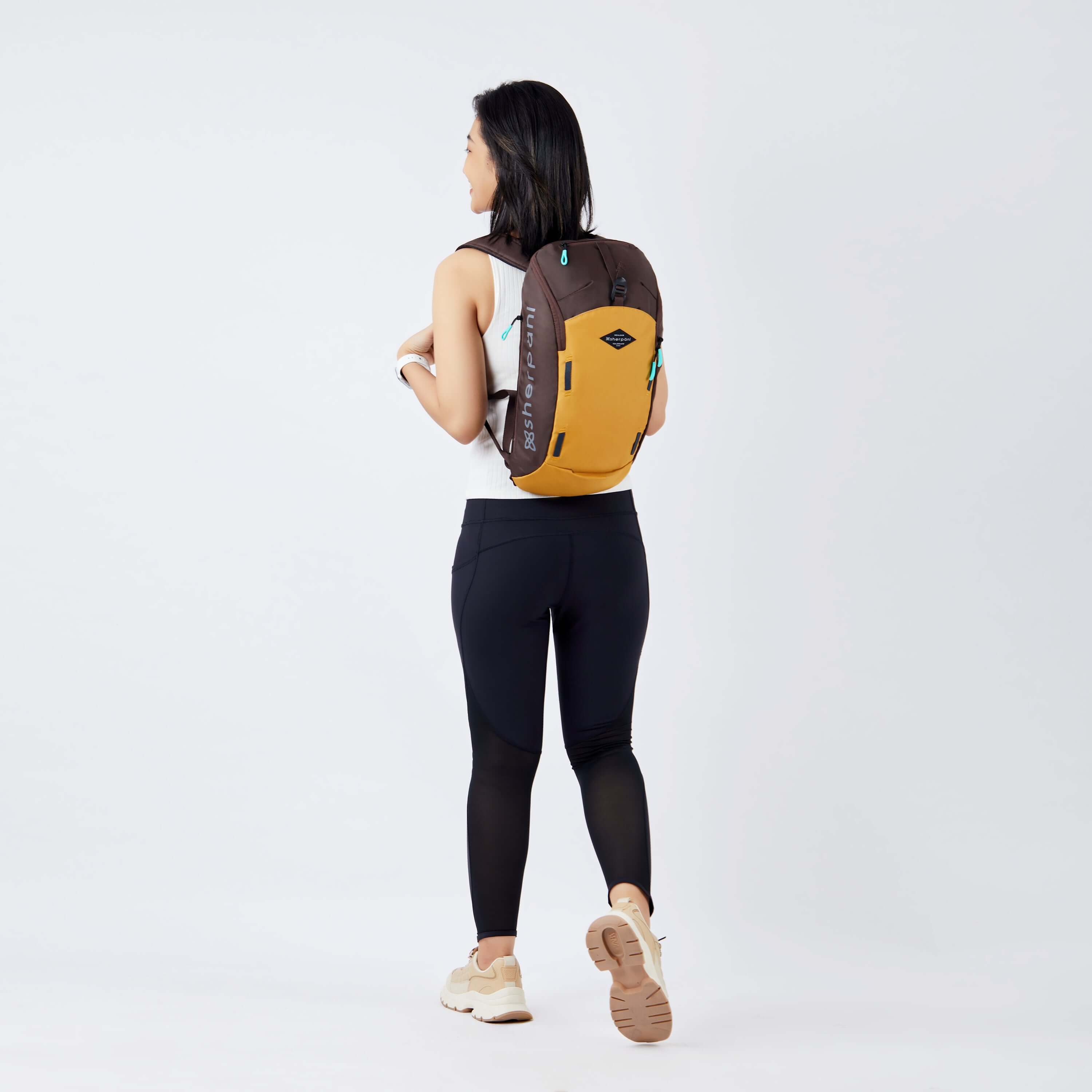 Full body view of a dark haired model facing away from the camera and walking. She is wearing a white tank top, black leggings and sneakers. She carries Sherpani backpack, the Switch in Sundial, on her shoulders. 