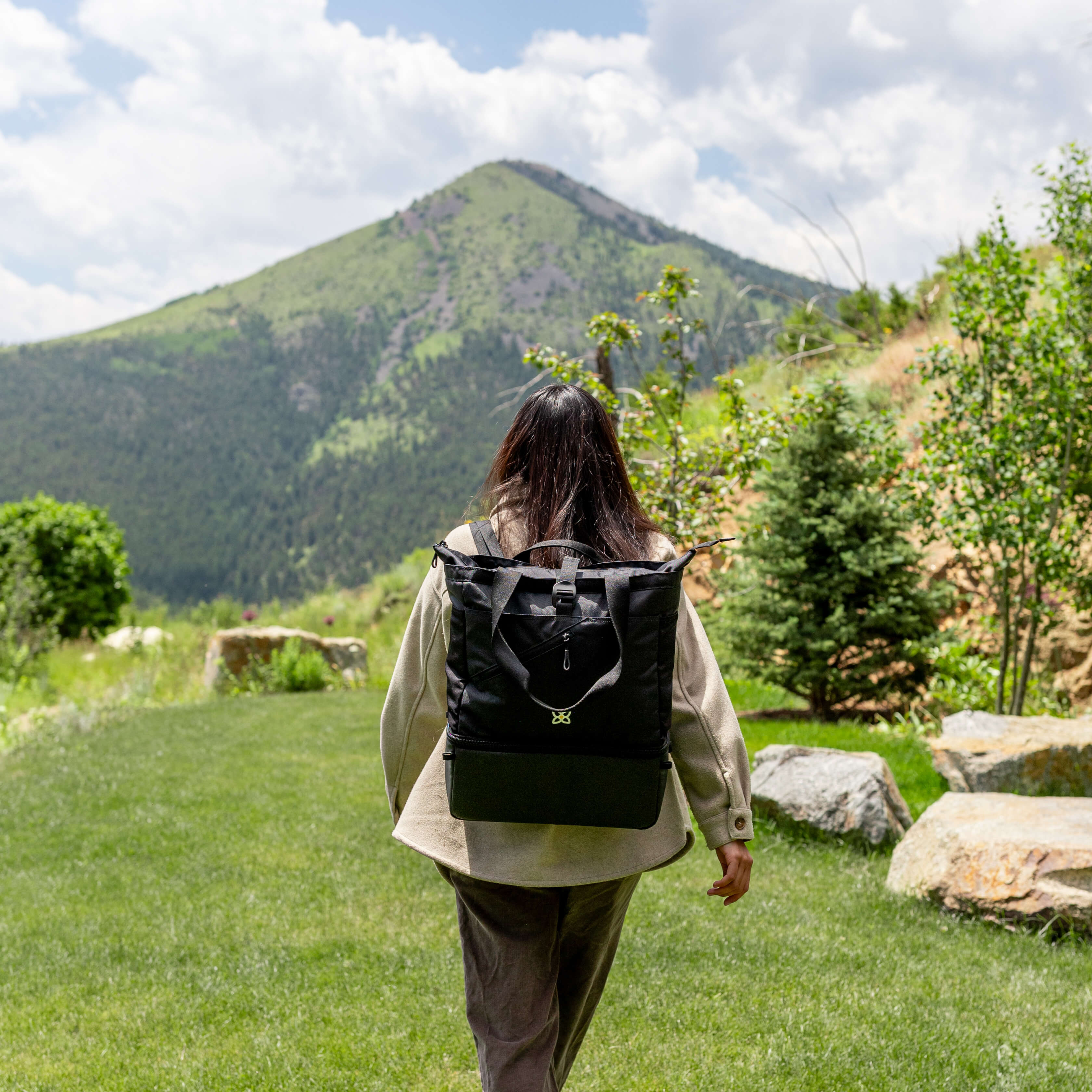 A woman walks outside in a scenic place with mountains, she is wearing Sherpani 2-in-1 tote/backpack combo, the Terra in Raven. 