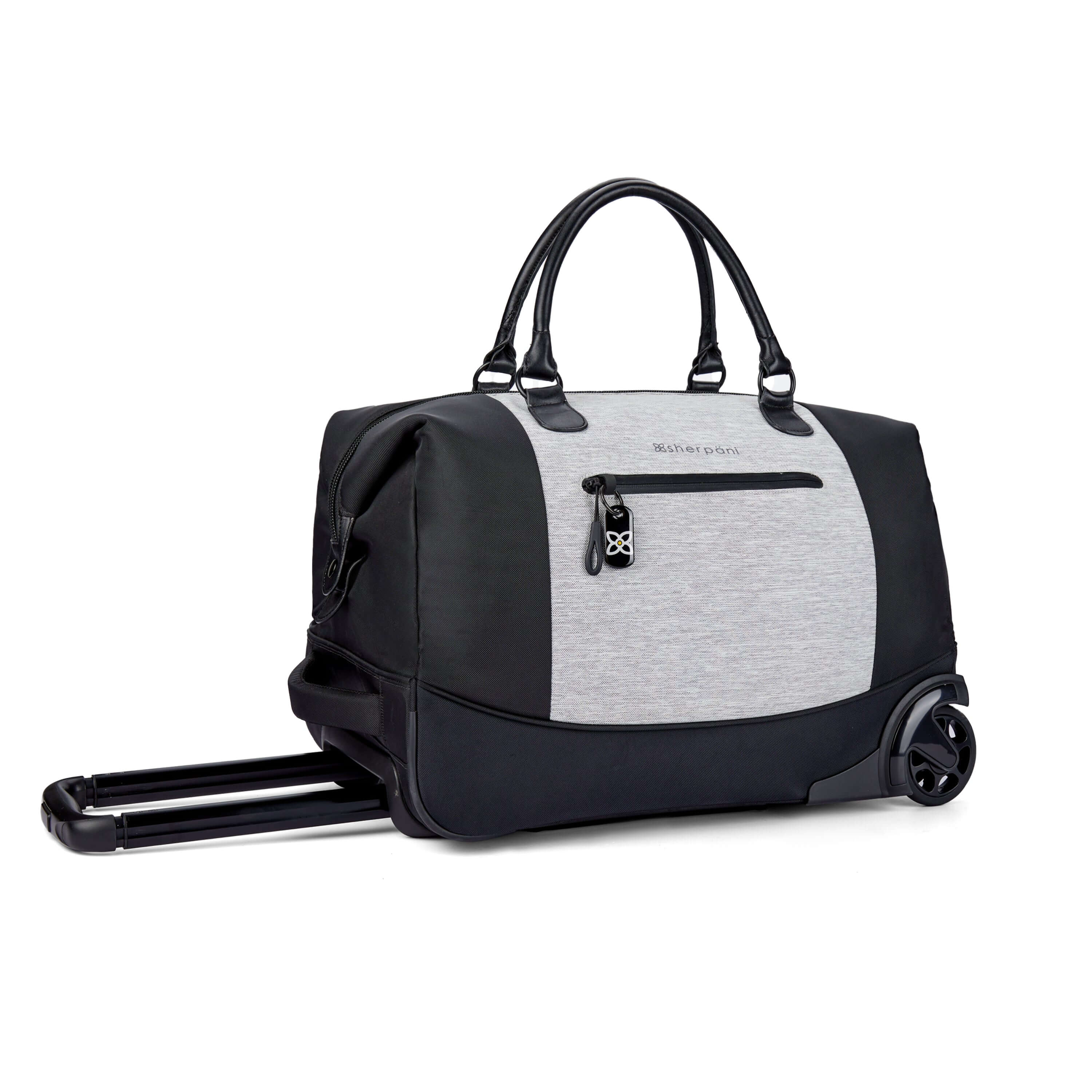 Angled front view of Sherpani’s Anti Theft rolling Duffle, the Trip in Sterling, with vegan leather accents in black. The bag lies flat on the ground with the luggage handle extended on the left side and the rolling wheels shown on the right side. The bag features short tote handles at the top and an external zipper compartment on the front panel with locking zipper and ReturnMe tag.