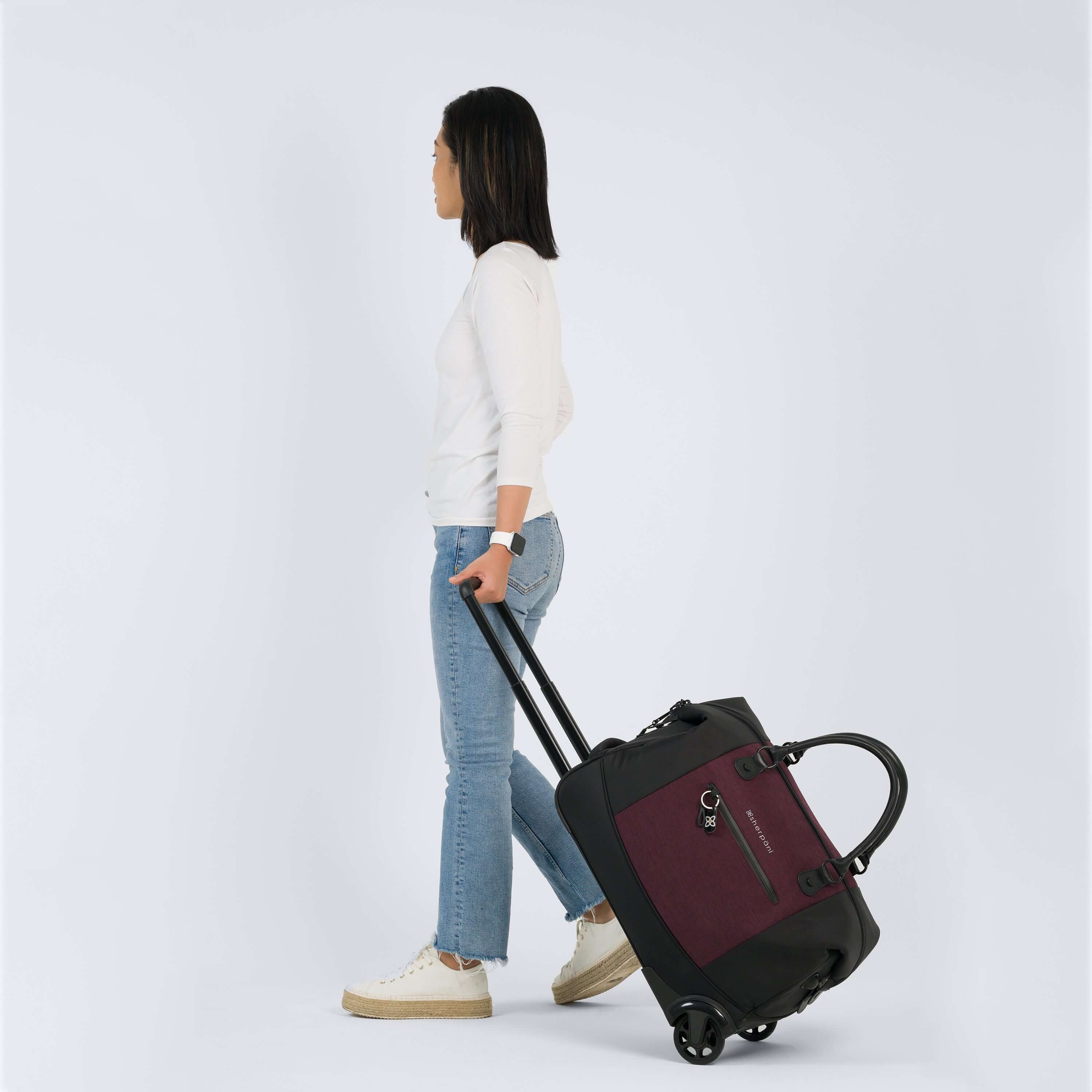 Full body view of a dark haired model facing the side. She is wearing a white shirt, jeans and white shoes. She is grabbing the luggage handle of Sherpani's Anti-Theft rolling duffle the Trip in Merlot, the bag rolls along beside her. 
