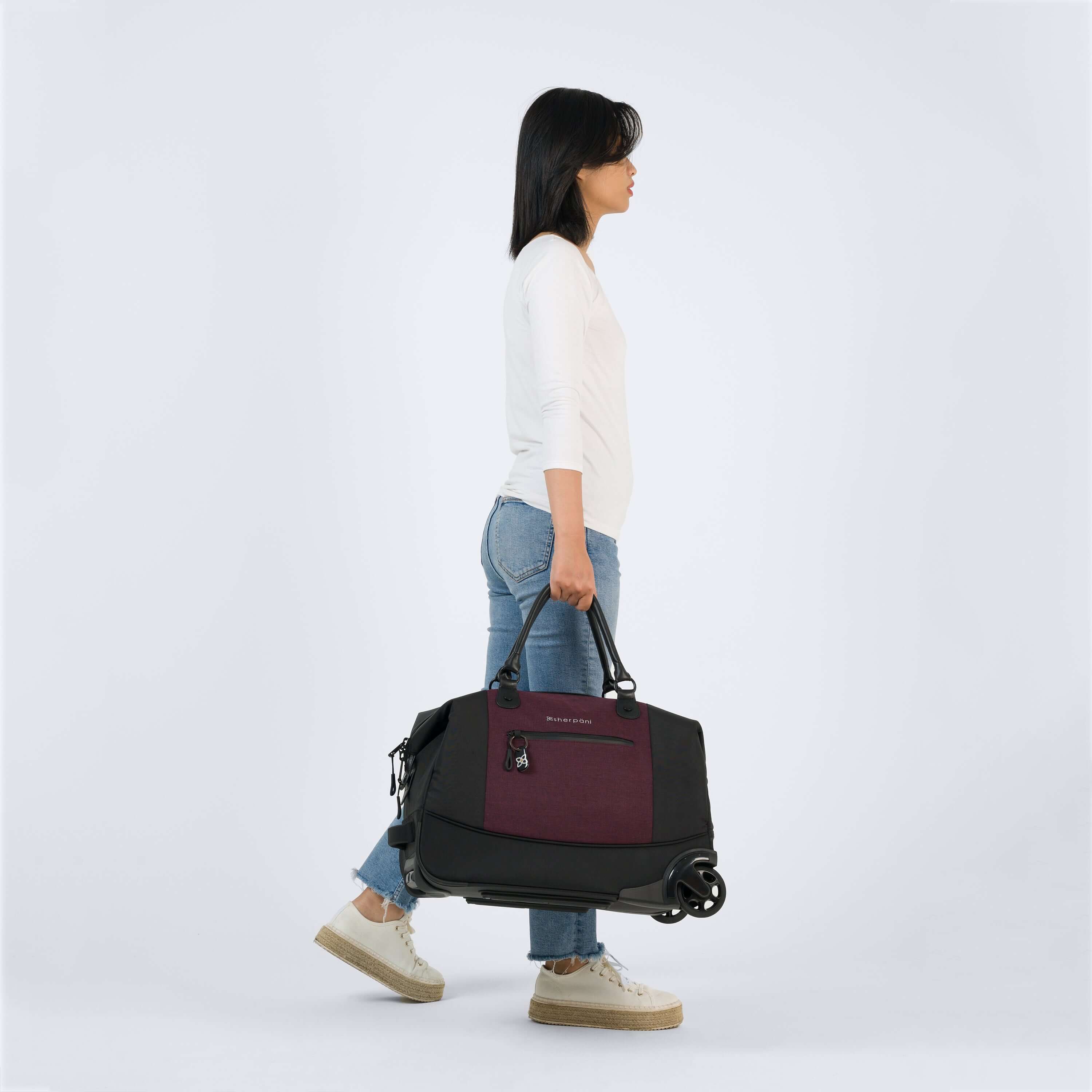 Full body view of a dark haired model facing the side. She is wearing a white shirt and blue jeans with white shoes. She is carrying Sherpani&#39;s Anti-Theft rolling duffle the Trip in Merlot by the tote handles.