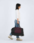 Full body view of a dark haired model facing the side. She is wearing a white shirt and blue jeans with white shoes. She is carrying Sherpani's Anti-Theft rolling duffle the Trip in Merlot by the tote handles.