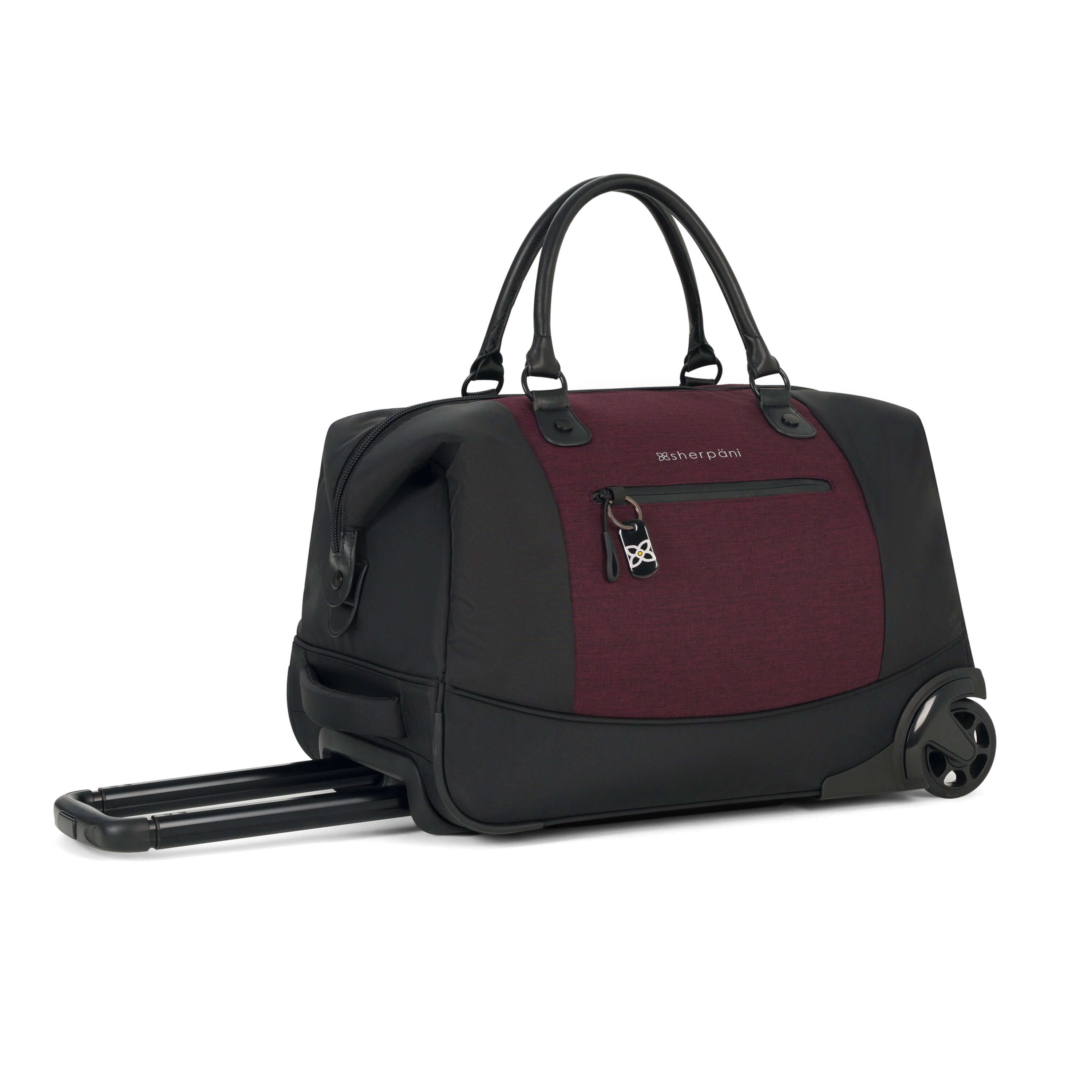 Angled front view of Sherpani’s Anti Theft rolling Duffle, the Trip in Merlot, with vegan leather accents in black. The bag lies flat on the ground with the luggage handle extended on the left side and the rolling wheels shown on the right side. The bag features short tote handles at the top and an external zipper compartment on the front panel with locking zipper and ReturnMe tag. #color_merlot