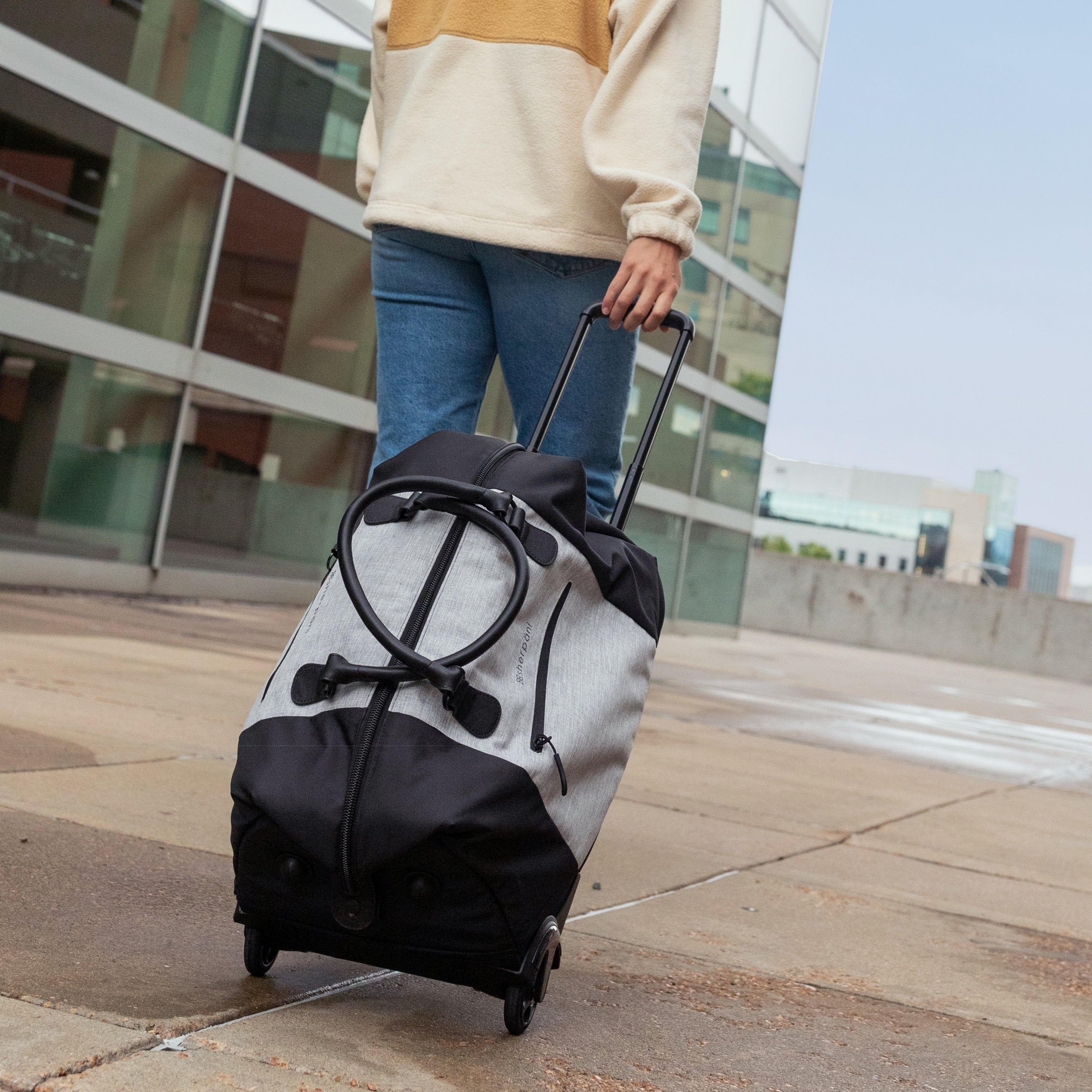 Close up view of a woman walking outside the airport. She is holding the luggage handle of the Trip in Sterling and pulling the large capacity rolling duffle bag along behind her. 