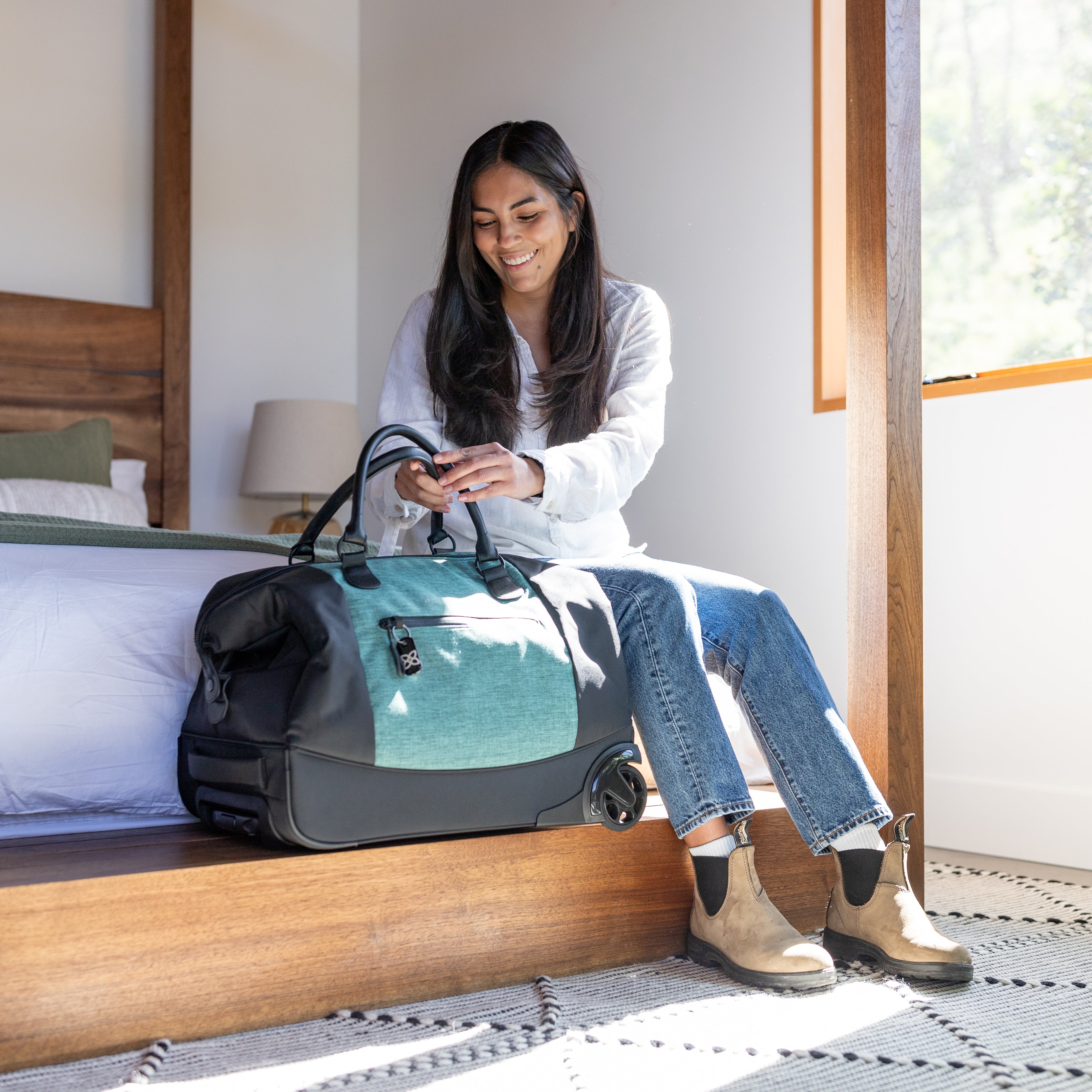 A woman sits on the bed in her hotel room. Next to her, is Sherpani Anti-Theft rolling duffle bag, the Trip in Teal. She is grabbing the attached tote bag handles.
