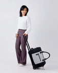 Full body view of a dark haired model facing the camera and smiling. She is wearing a white shirt and purple leggings. She is holding the luggage handle of Sherpani's Anti-Theft rolling duffle the Trip in Sterling.