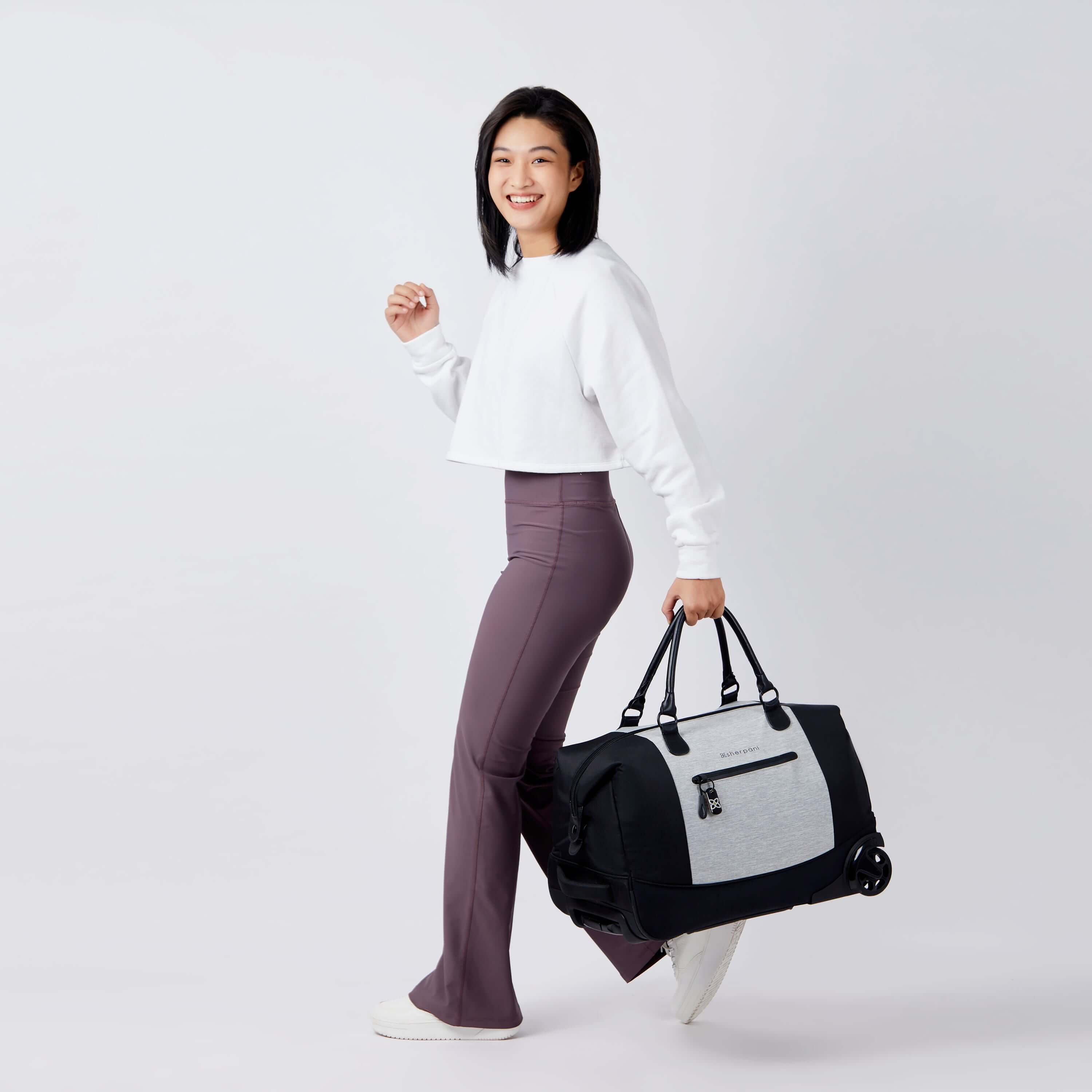 Full bodied view of a dark haired model facing the side and smiling at the camera. She is wearing a white shirt and purple leggings. She carries Sherpani's Anti-Theft rolling duffle the Trip in Sterling by the tote handles. 