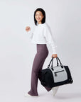 Full bodied view of a dark haired model facing the side and smiling at the camera. She is wearing a white shirt and purple leggings. She carries Sherpani's Anti-Theft rolling duffle the Trip in Sterling by the tote handles.