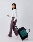 Full bodied view of a dark haired model facing the side and is smiling over her left shoulder at the camera. She is wearing a white shirt and purple leggings. She is holding the luggage handle of Sherpani's Anti-Theft rolling duffle the Trip in Teal as the bag rolls along beside her.