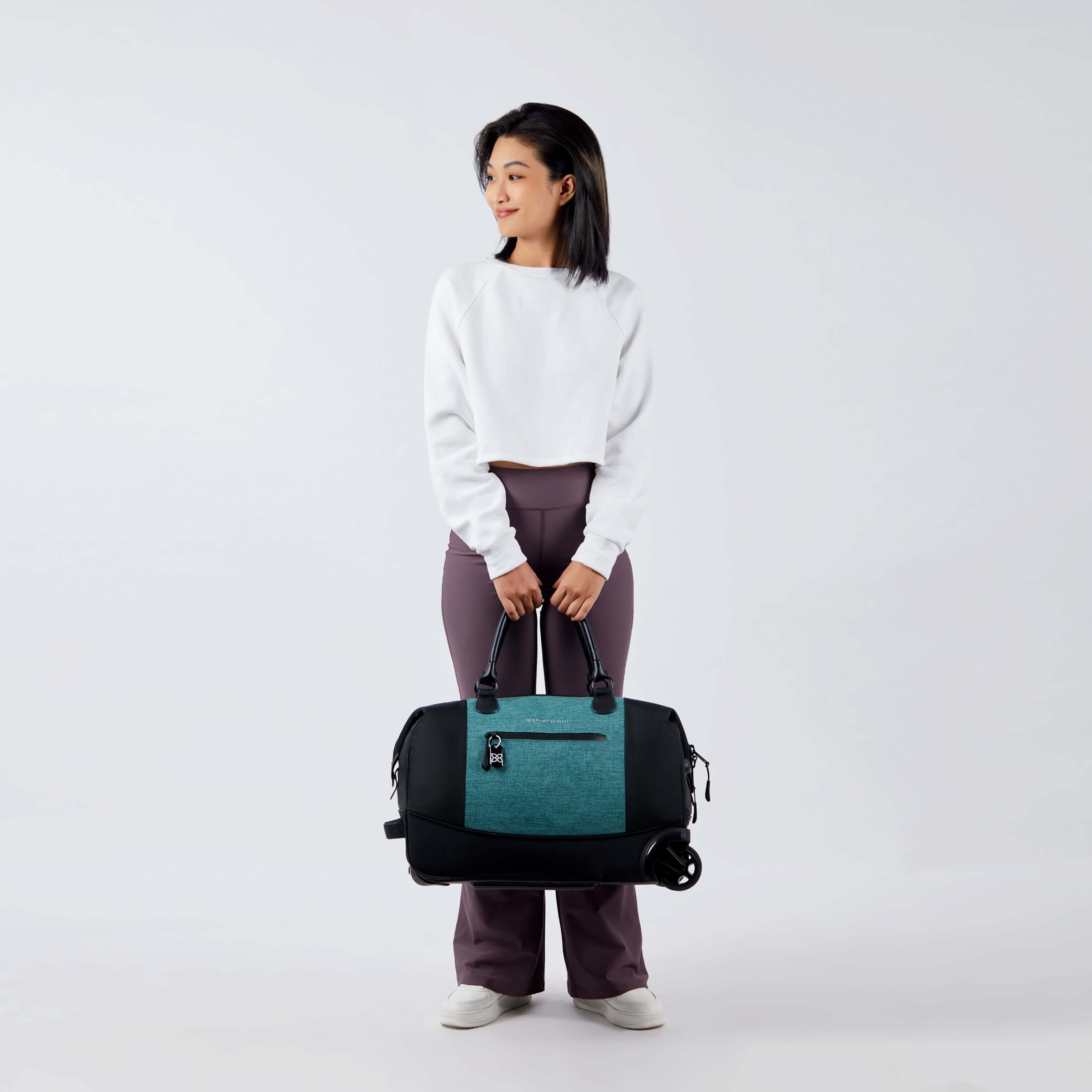 Full bodied view of a dark haired model facing the camera and smiling over her right shoulder. She is wearing a white shirt and purple leggings. She carries Sherpani's Anti-Theft rolling duffle the Trip in Teal by the tote handles. 