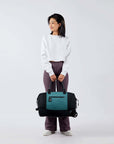 Full bodied view of a dark haired model facing the camera and smiling over her right shoulder. She is wearing a white shirt and purple leggings. She carries Sherpani's Anti-Theft rolling duffle the Trip in Teal by the tote handles.