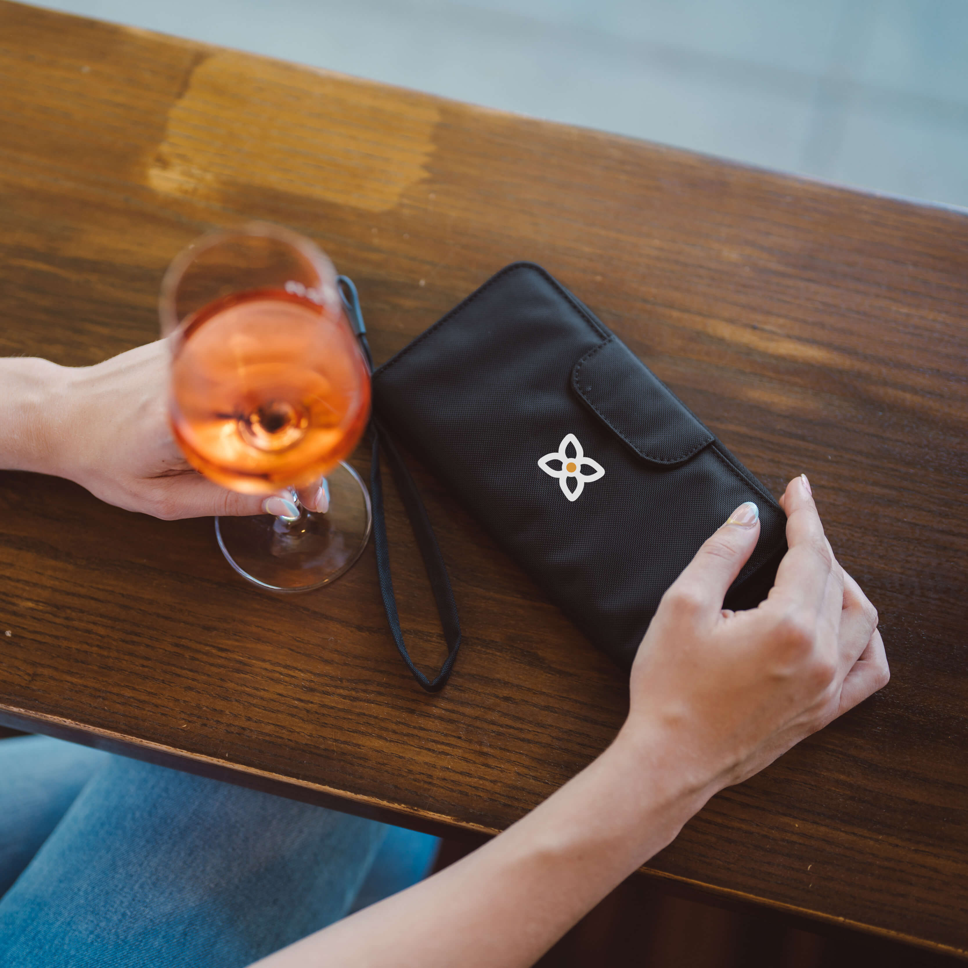 Close up view of a woman sitting at a table. On the table is a glass of wine and Sherpani RFID blocking wallet, the Tulum in Raven. 