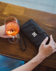 Close up view of a woman sitting at a table. On the table is a glass of wine and Sherpani RFID blocking wallet, the Tulum in Raven.
