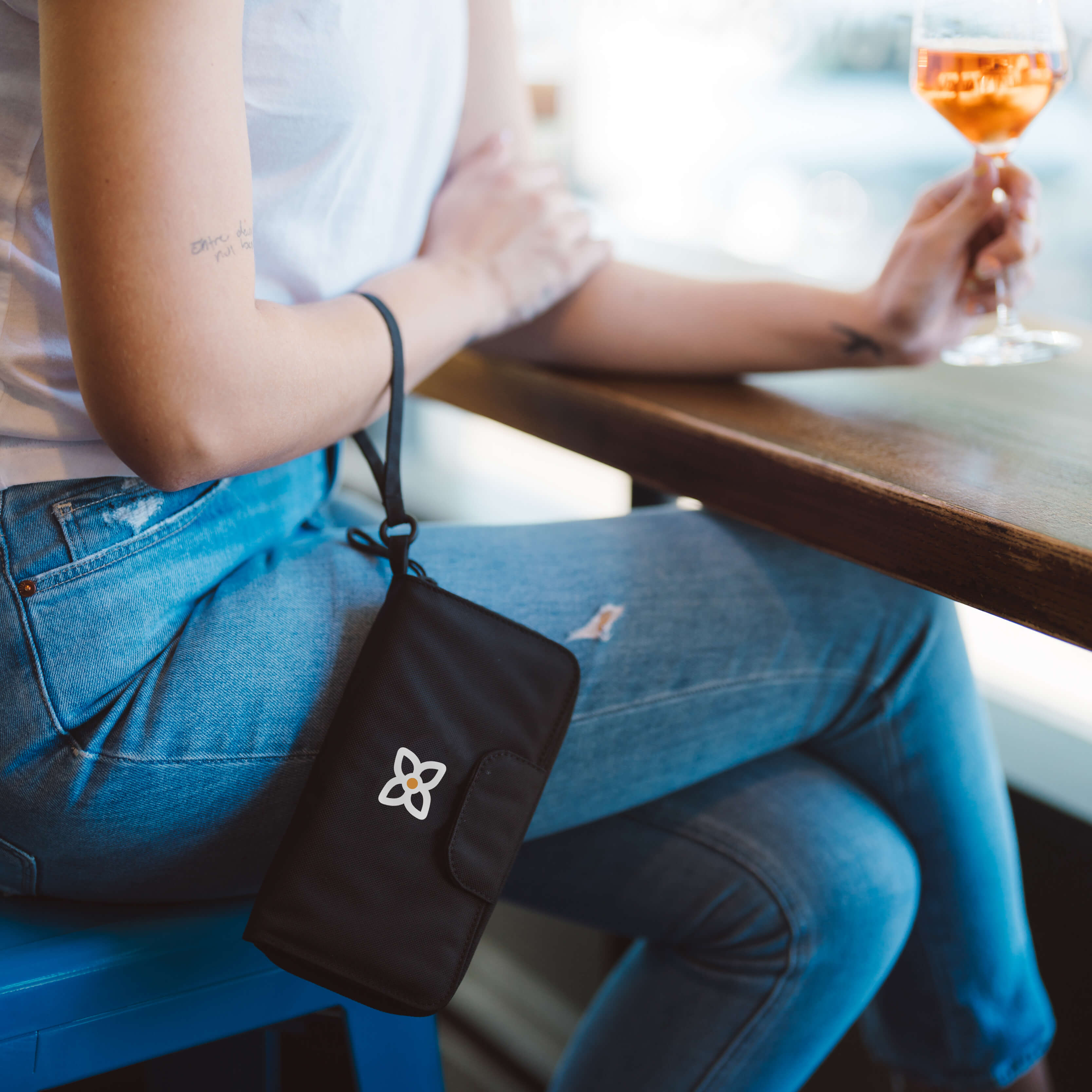 A woman sits at a table with a glass of wine. Dangling from her writs is Sherpani RFID blocking wallet for women, the Tulum in Raven. 