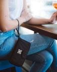 A woman sits at a table with a glass of wine. Dangling from her writs is Sherpani RFID blocking wallet for women, the Tulum in Raven.