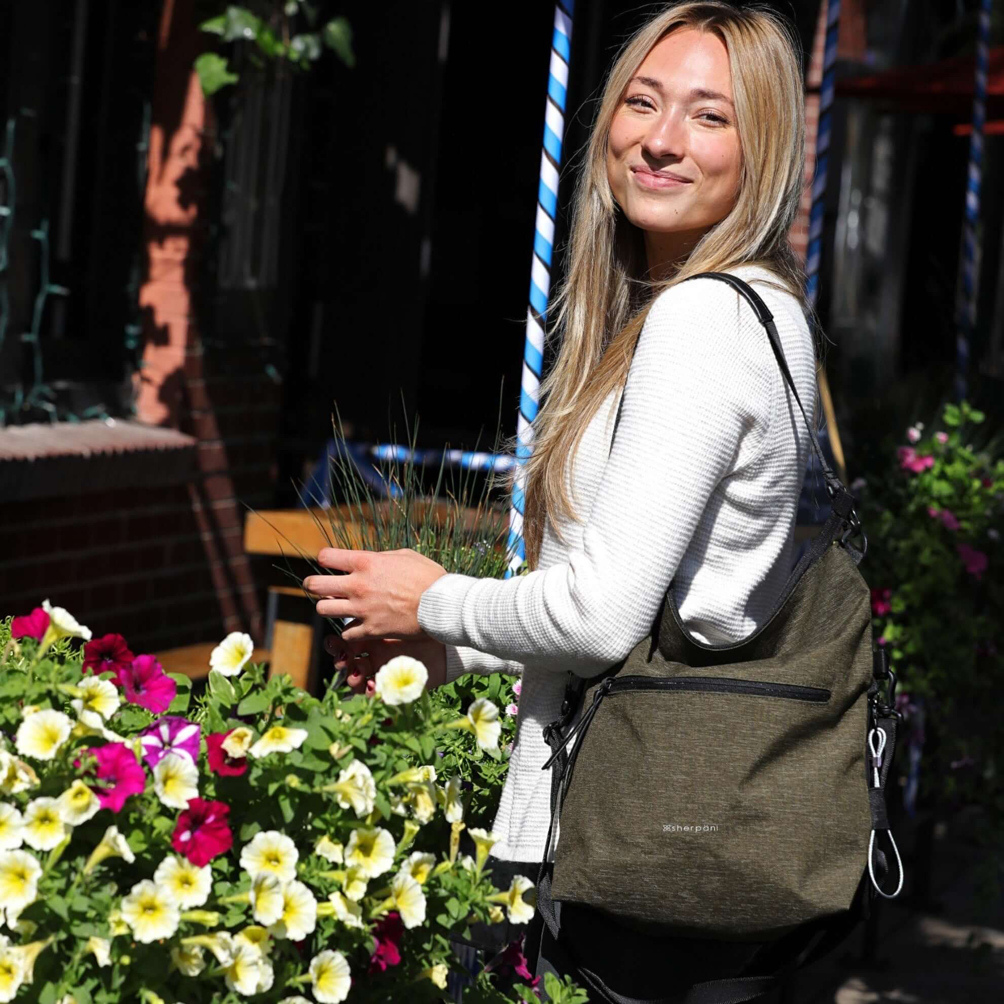 Close up view of a blonde model smiling over her left shoulder at the camera. She is wearing a long sleeved white shirt, and Sherpani&#39;s Anti-Theft bag, the Vale AT in Loden, as a tote.