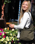 Close up view of a blonde model smiling over her left shoulder at the camera. She is wearing a long sleeved white shirt, and Sherpani's Anti-Theft bag, the Vale AT in Loden, as a tote.