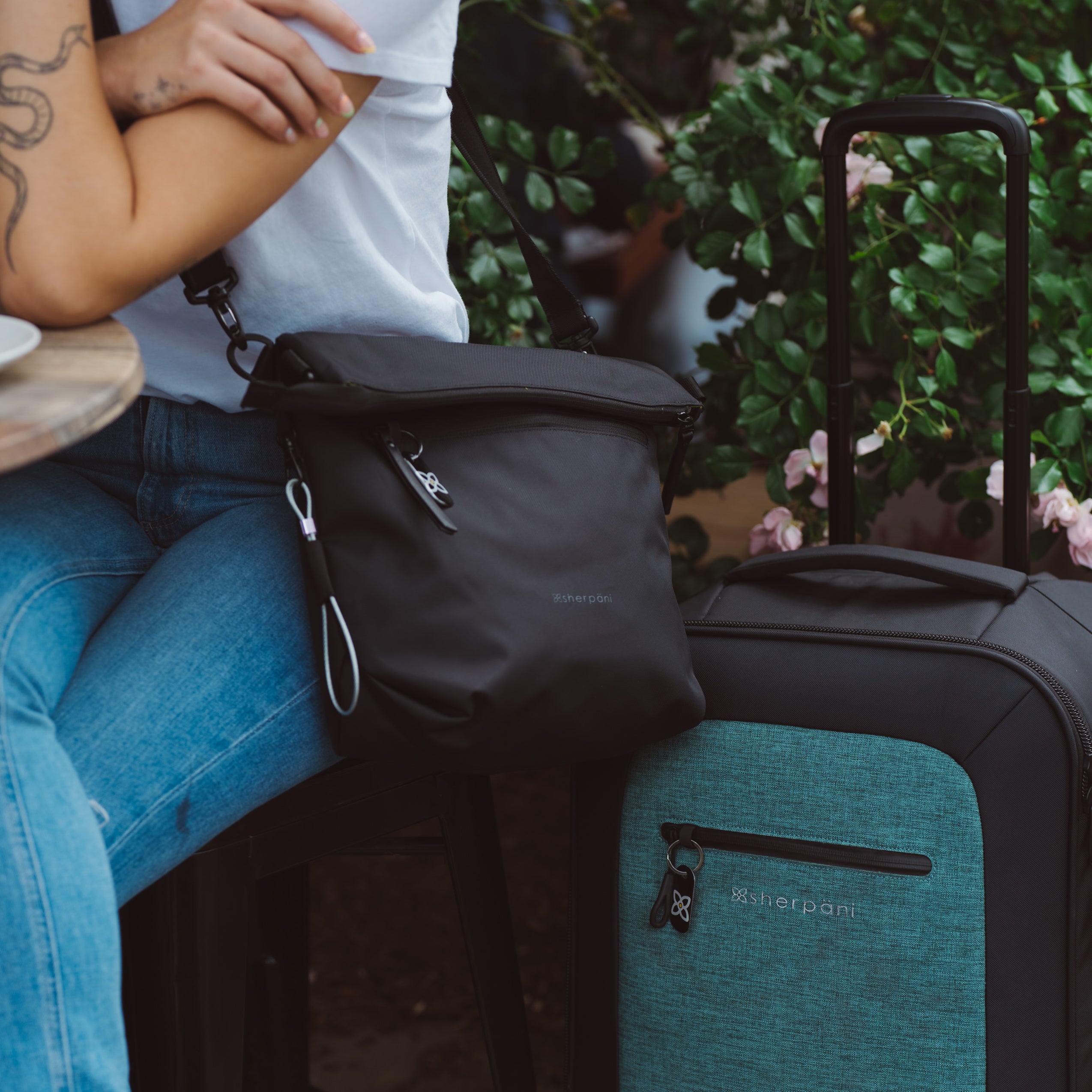 Close up view of a woman sitting outdoors wearing Sherpani reversible bag, the Vale in Carbon, as an Anti-Theft crossbody purse. Next to her sits Sherpani soft-shell carry-on luggage, the Latitude in Teal. 