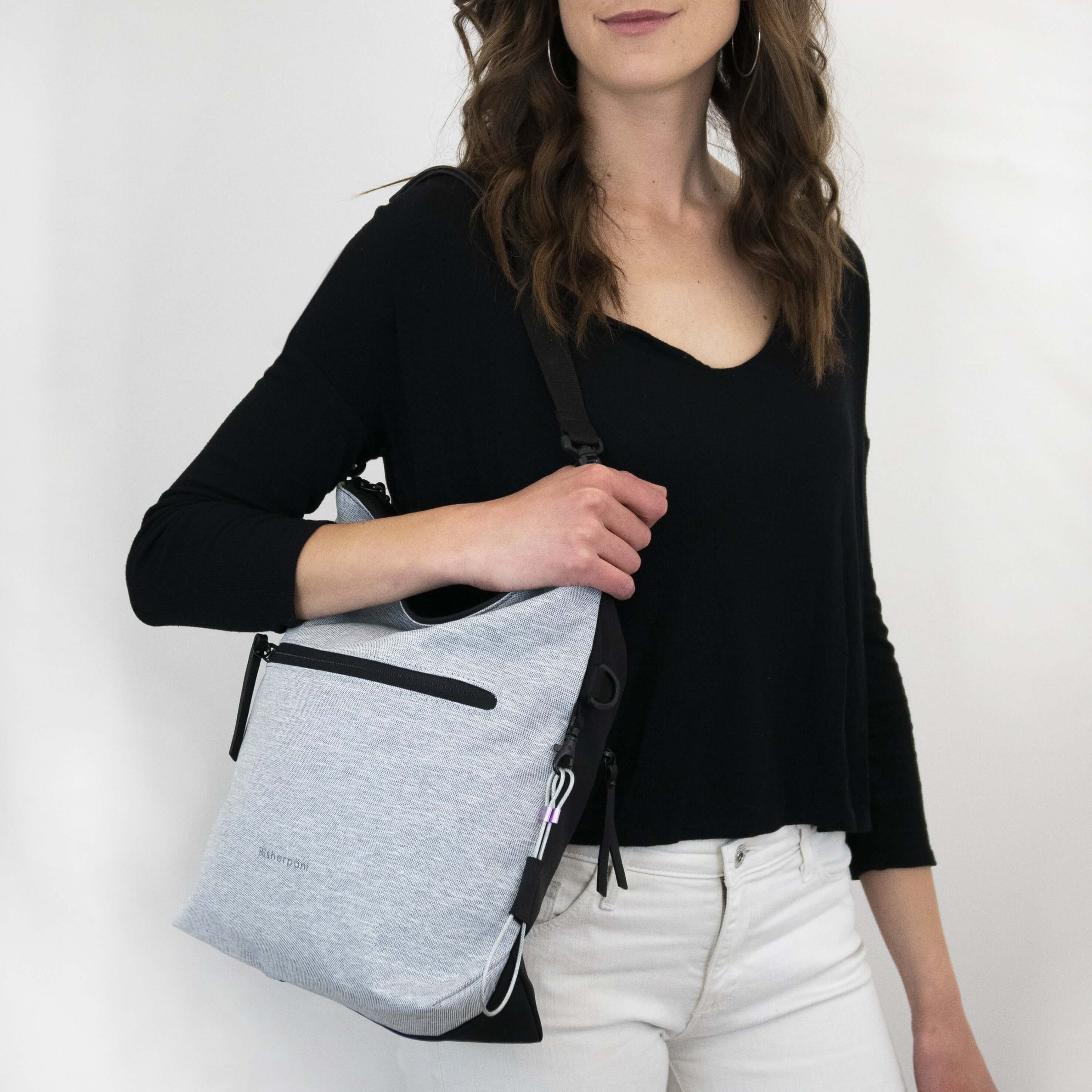 Close up view of a model facing the camera and smiling. She is wearing a black shirt and white pants. She carries Sherpani&#39;s Anti-Theft bag, the Vale AT in Sterling, over her shoulder as a tote.