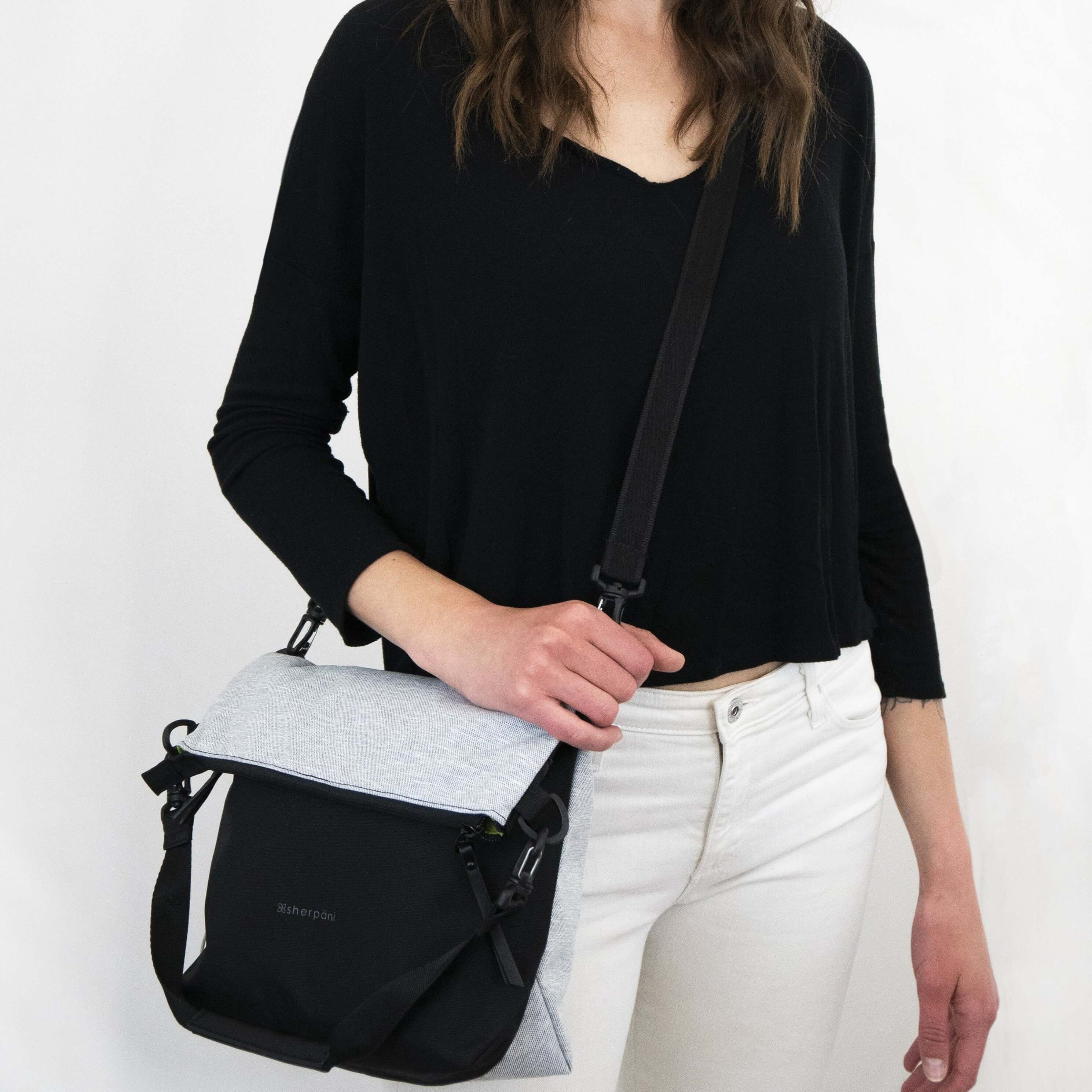 Close up view of brown haired model facing the camera. She is wearing a black shirt, white pants, and Sherpani&#39;s Anti-Theft bag, the Vale AT in Sterling, as a crossbody.