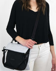 Close up view of brown haired model facing the camera. She is wearing a black shirt, white pants, and Sherpani's Anti-Theft bag, the Vale AT in Sterling, as a crossbody.