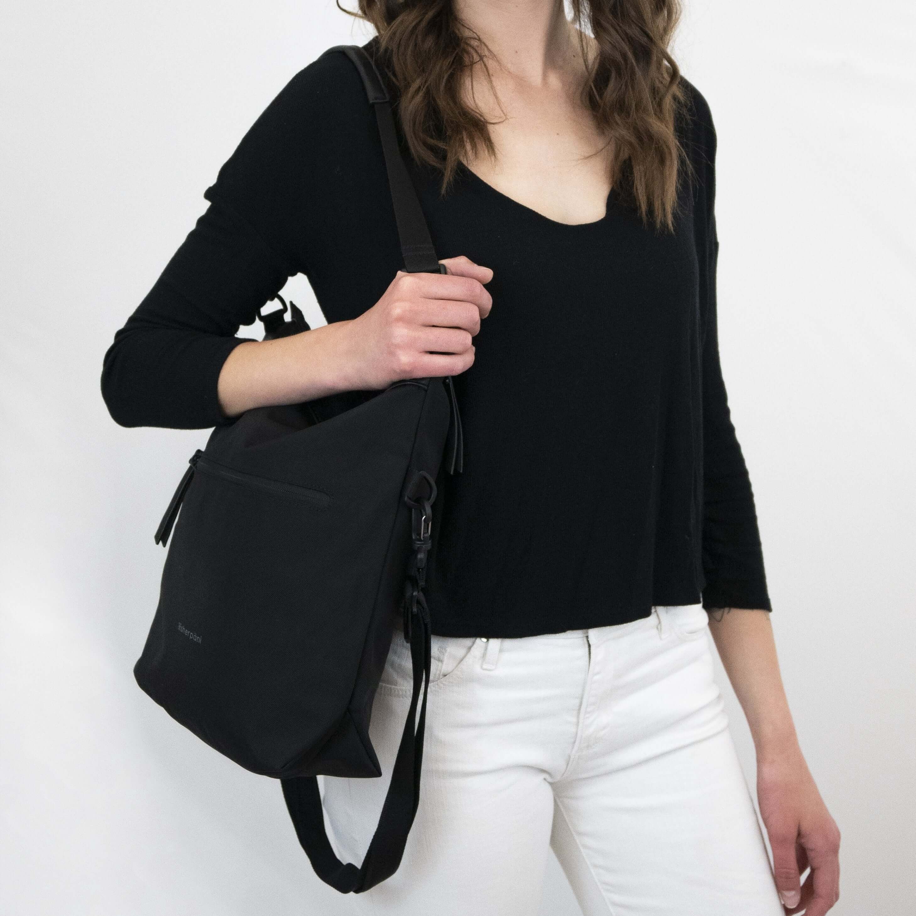 Close up view of a brown haired model facing the camera. She is wearing a black shirt and white pants. She carries Sherpani&#39;s Anti-Theft bag, the Vale AT in Carbon, over her shoulder as a tote.